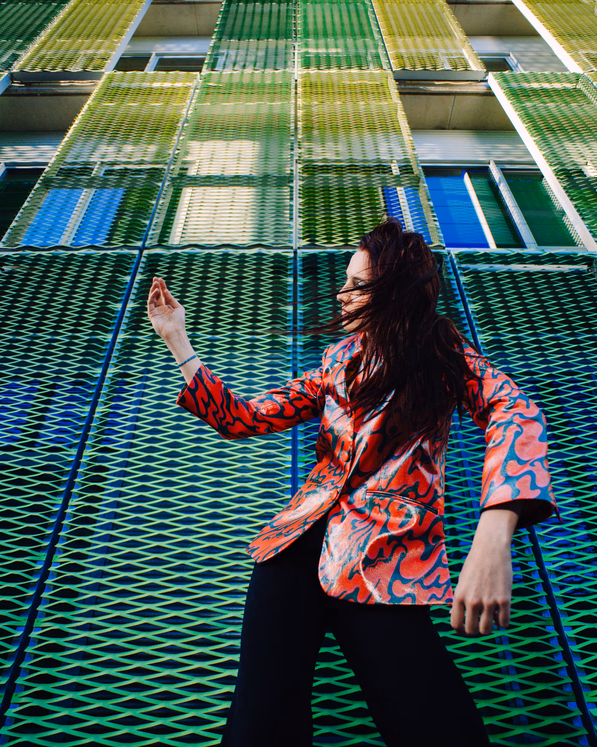 Woman with long dark hair wearing a colorful red and blue patterned jacket, posed dynamically against a building facade with green metal mesh panels.