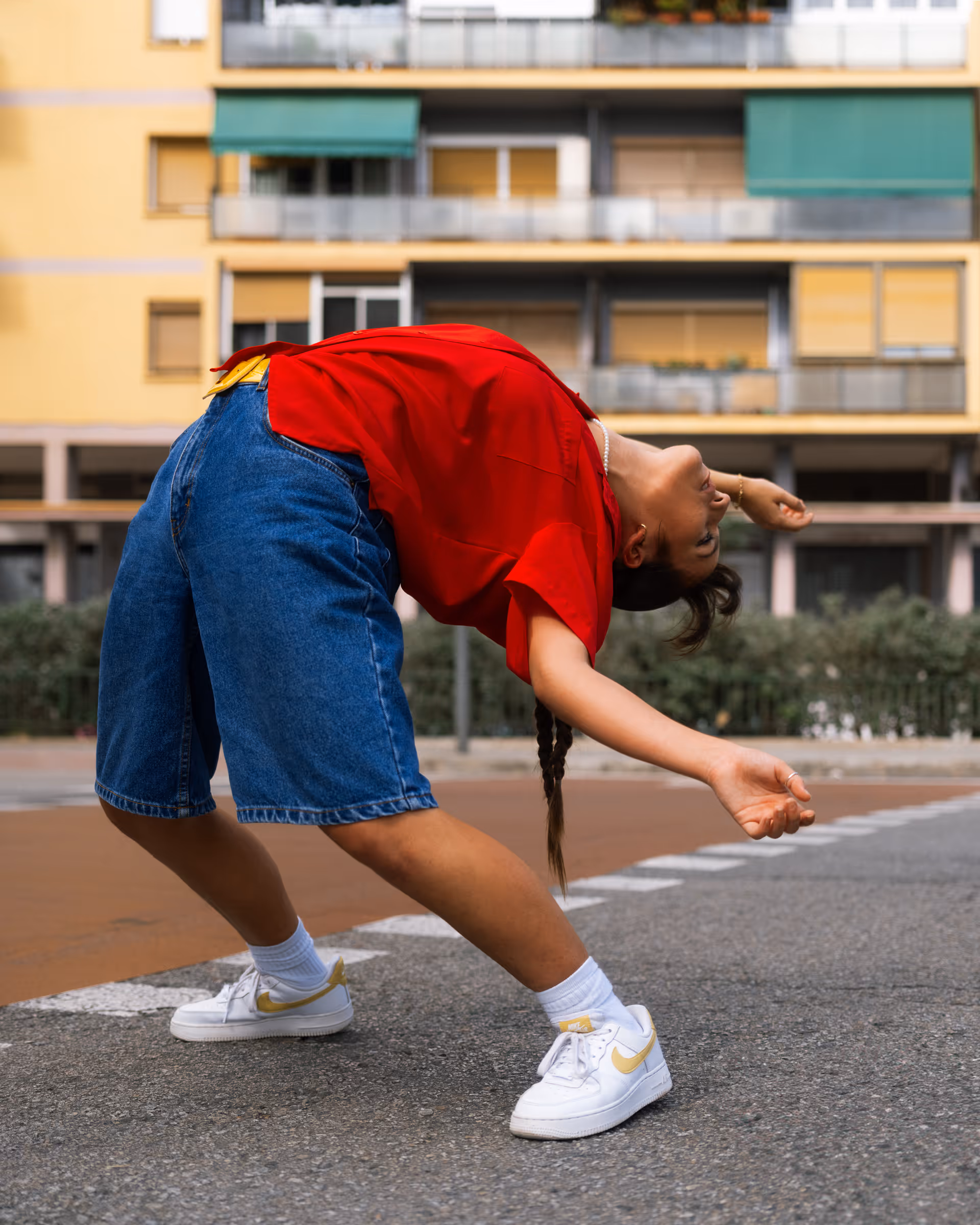Person wearing a red shirt and blue shorts bending backward in a dance pose on a city street.