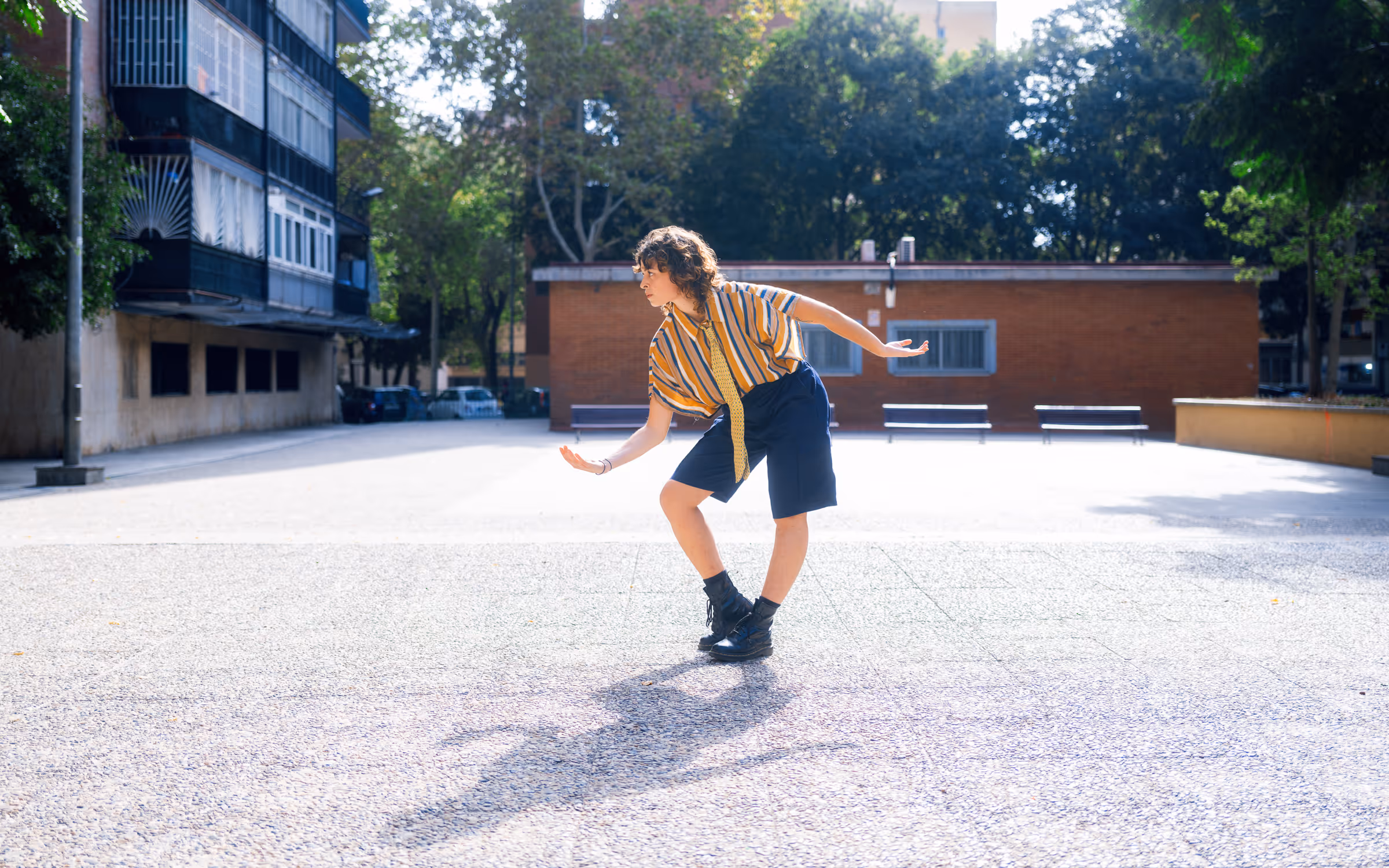 Person in striped shirt and shorts balancing in a playful pose on a sunlit urban plaza.