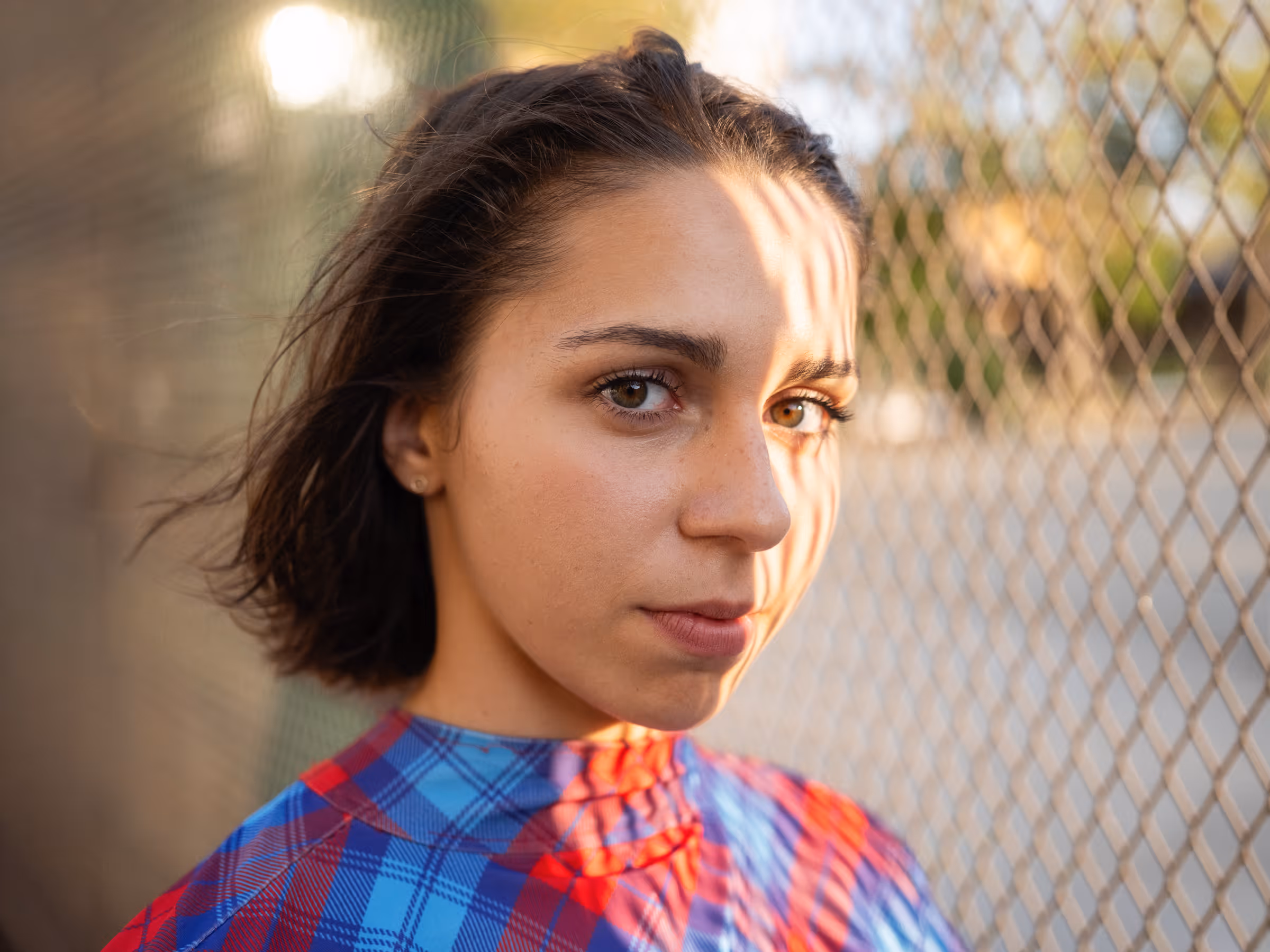 Close-up of a woman with short dark hair wearing a blue and red plaid shirt standing in front of a chain-link fence with sunlight casting shadows on her face.