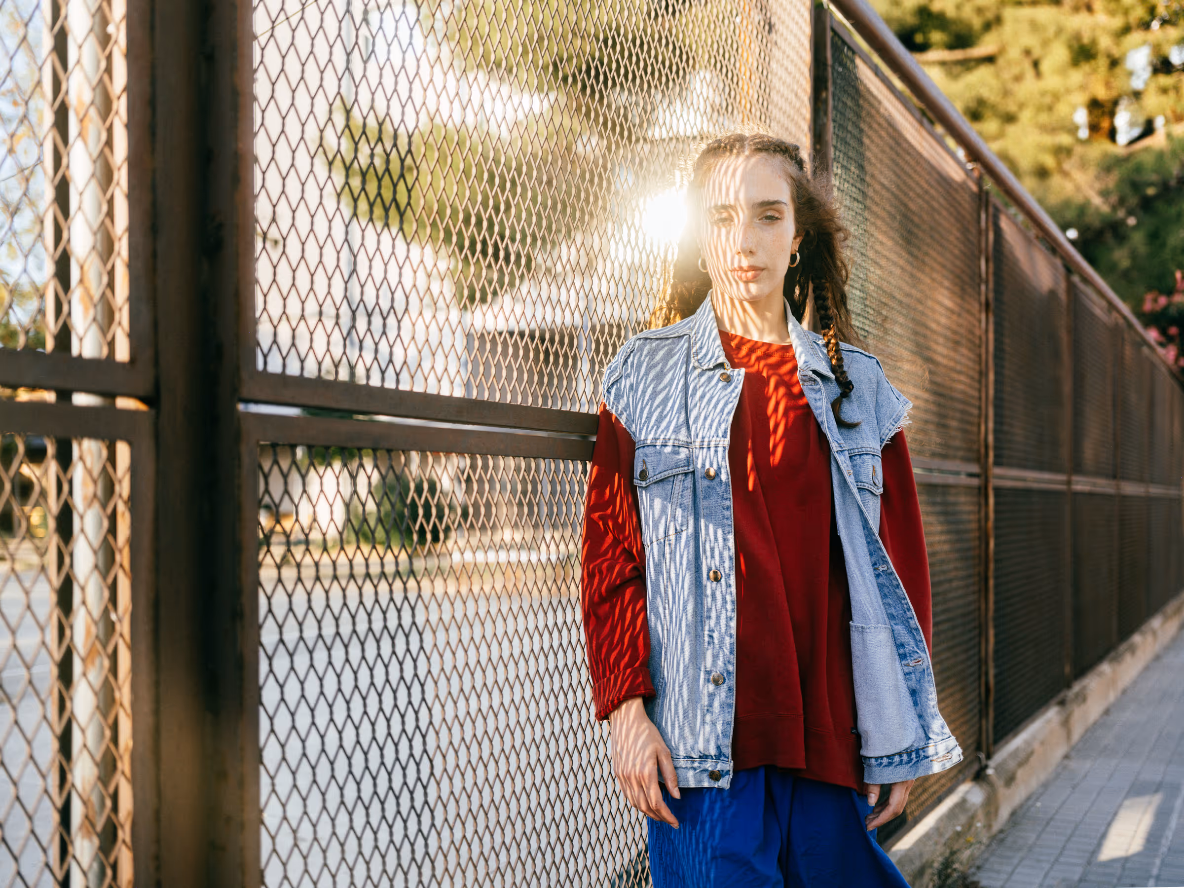 Young woman with braids wearing a red long-sleeve shirt, light denim vest, and blue pants standing by a metal fence with sunlight casting patterned shadows on her face and clothes.