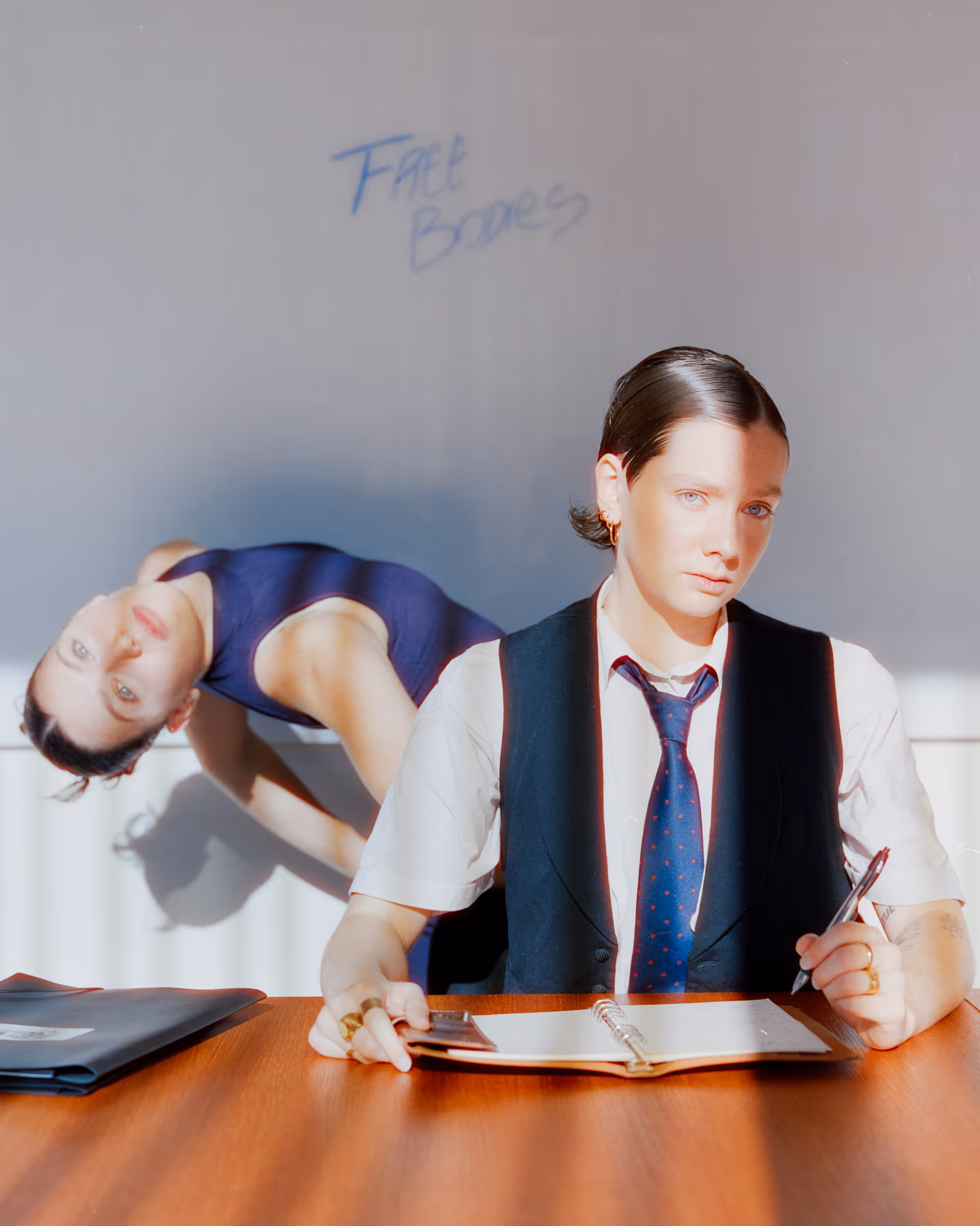 Person in a white shirt, black vest, and blue tie sitting at a table with an open notebook, while another person in a blue sleeveless outfit bends sideways behind them, with 'Free Bodies' written on the wall.
