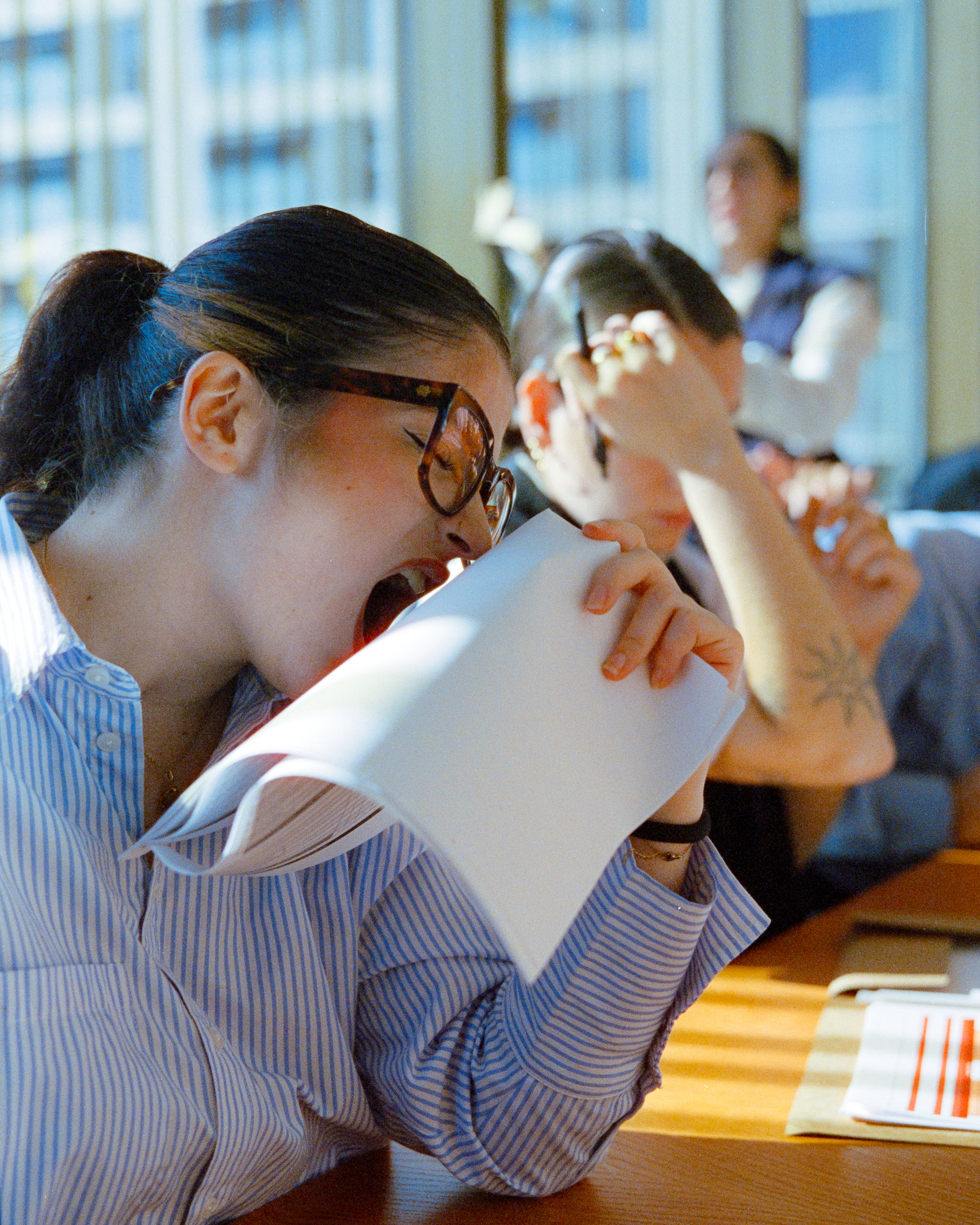 Woman wearing glasses and a striped shirt biting into a notebook paper in a bright room with others in the background.
