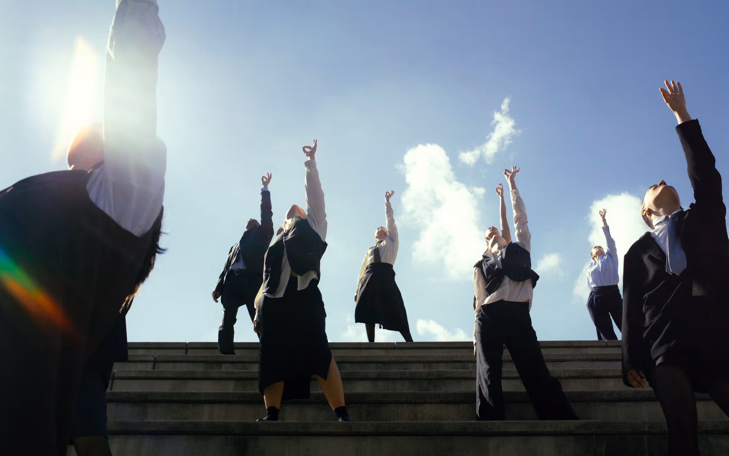 Group of people dressed in business attire standing on stairs reaching one arm upward against a blue sky with clouds.