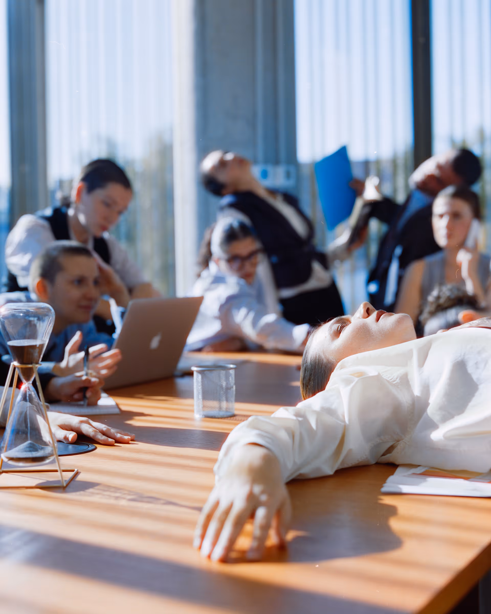 Tired businesswoman lying on a conference table surrounded by coworkers appearing stressed and overwhelmed during a meeting.