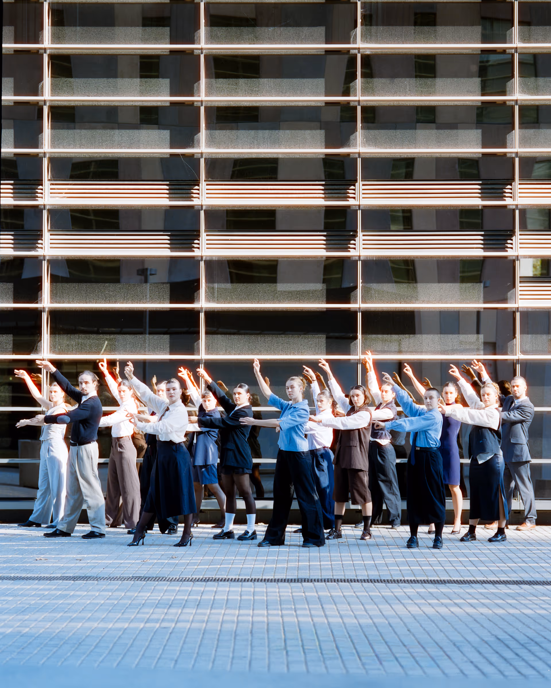 A group of people standing in a line outdoors in front of a reflective glass building, posing with one arm extended forward and the other raised above their heads.