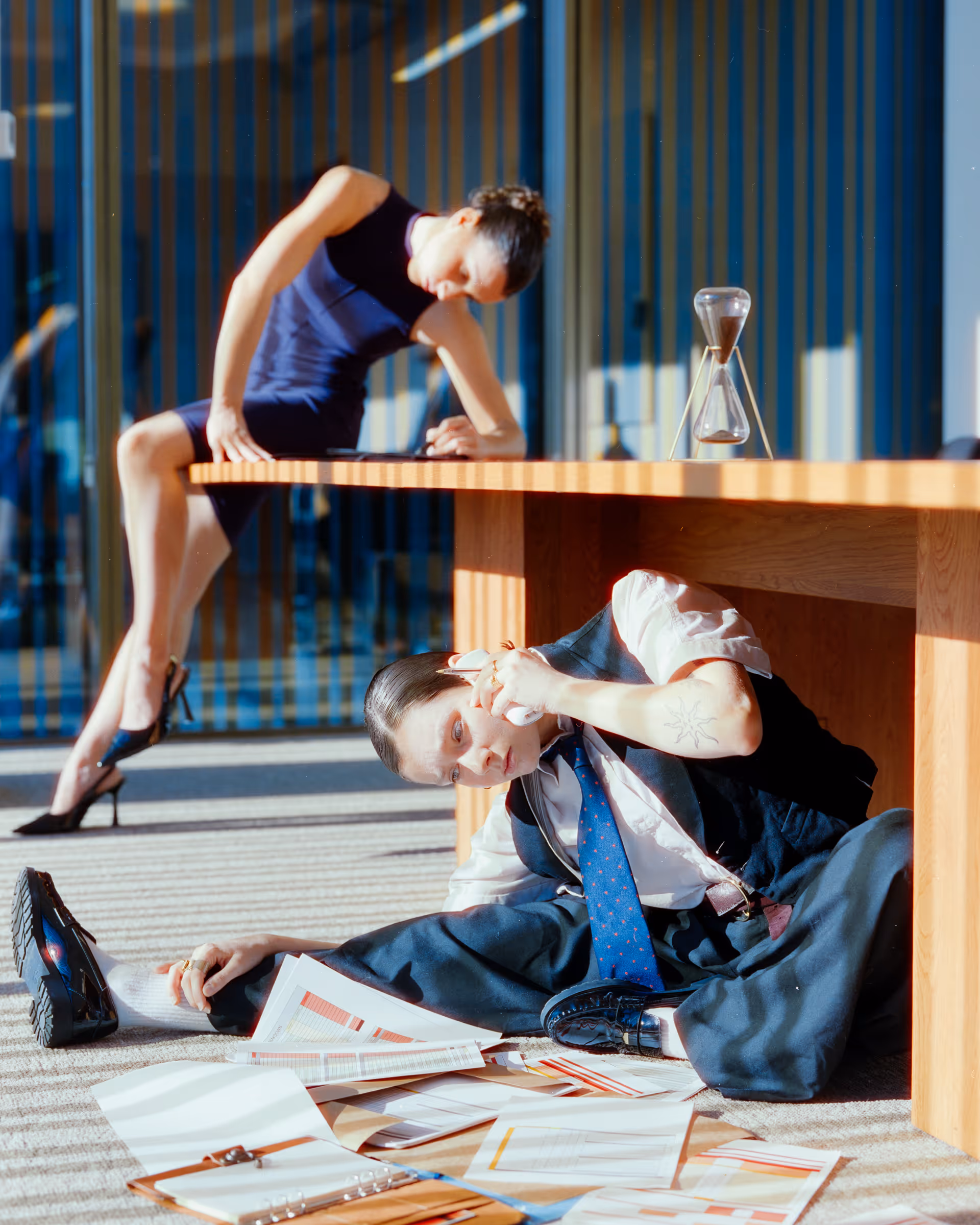 Person in formal attire sitting on the floor under a wooden table surrounded by scattered documents, talking on a phone, with another person sitting on the table in the background.
