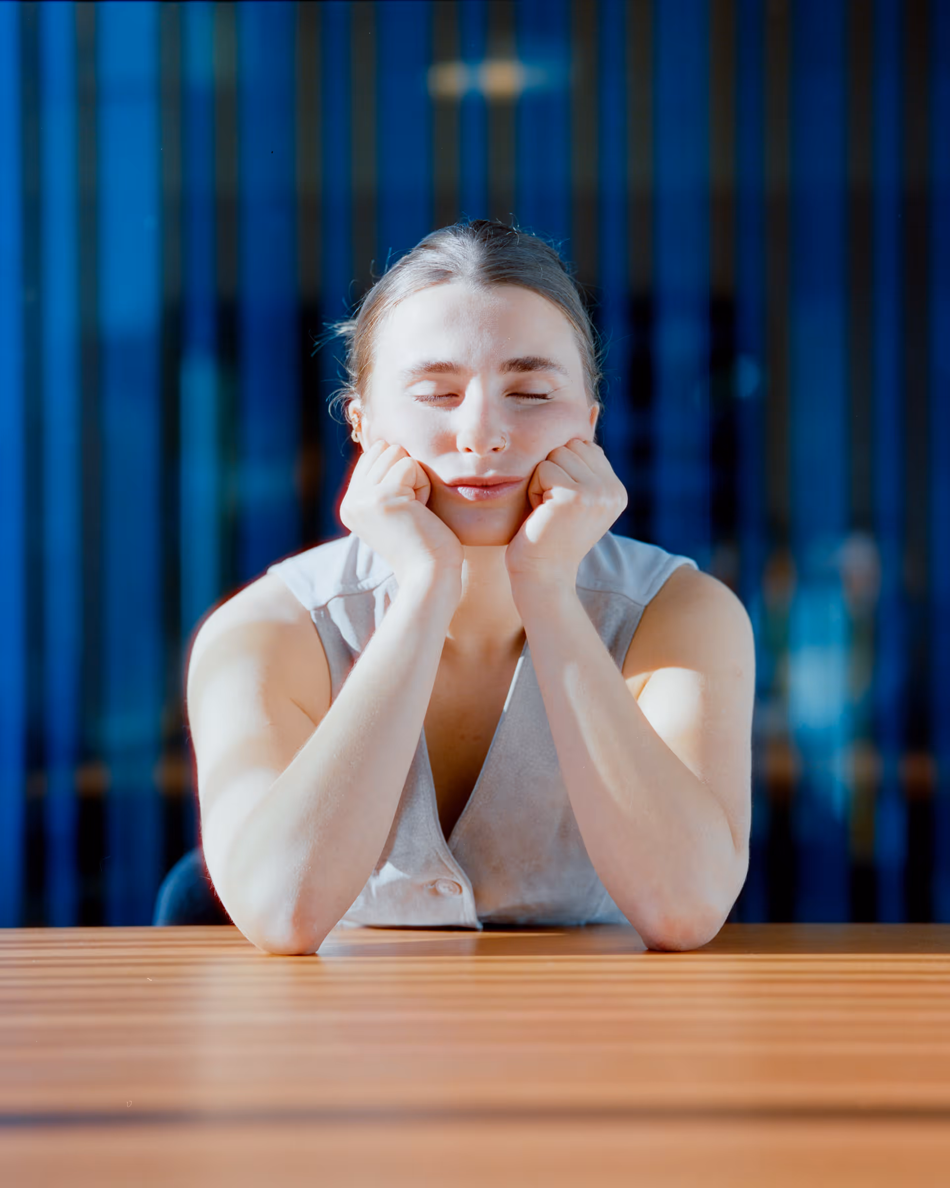 Young woman with eyes closed resting her chin in her hands on a wooden table, with a blue striped background.