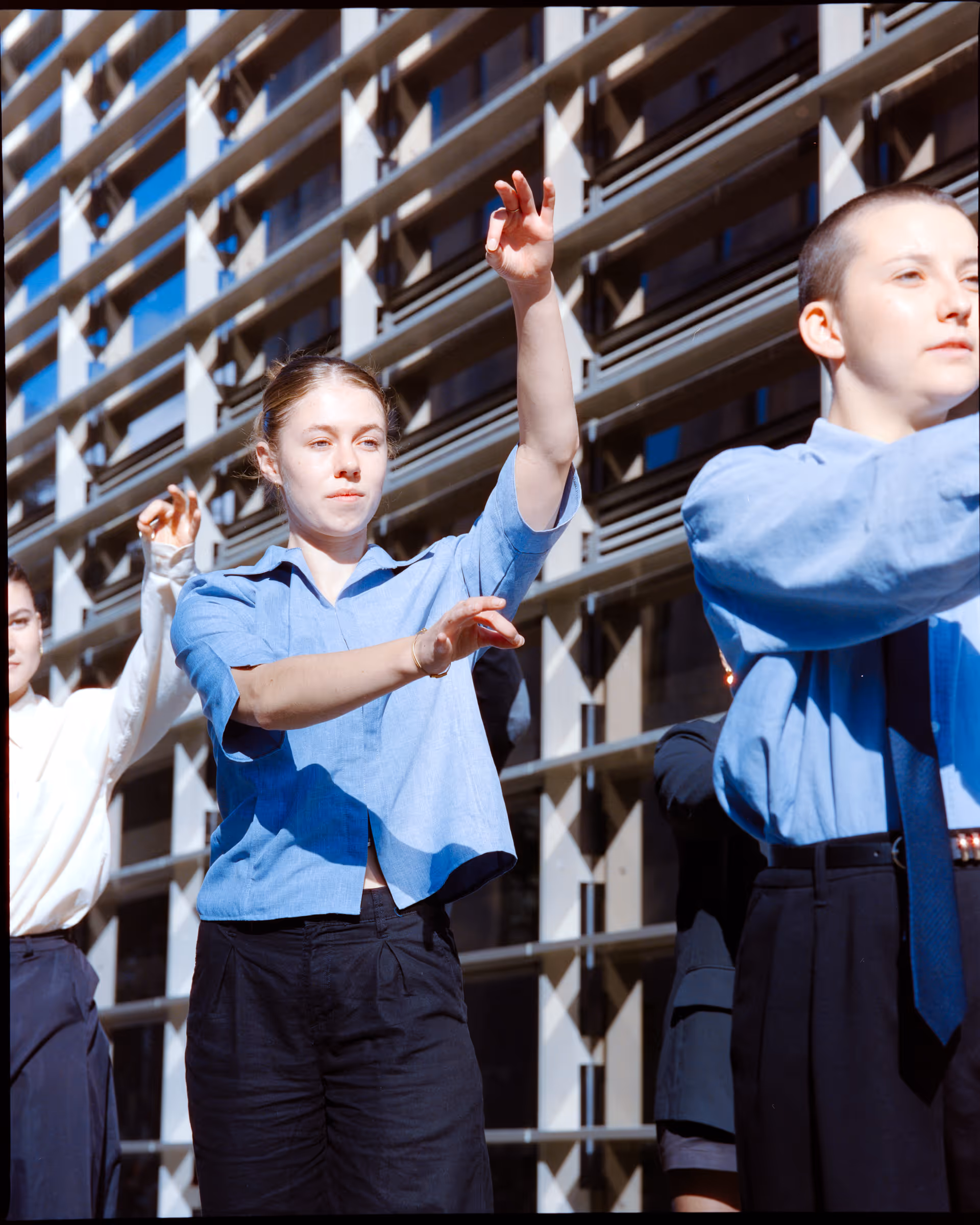 Young adults in blue and white shirts performing synchronized hand movements outside near a modern building facade.
