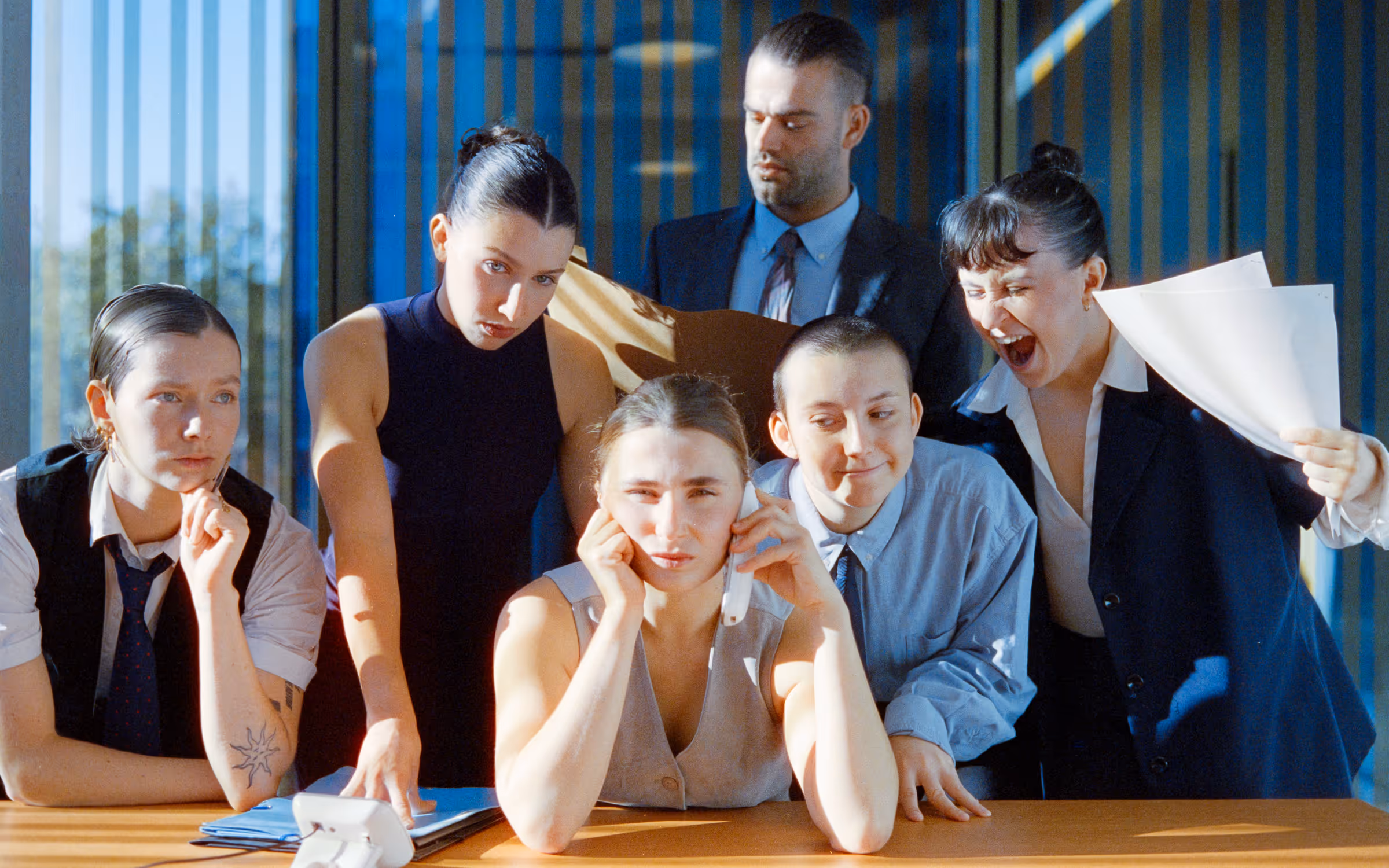 Five coworkers in an office; one woman looks stressed on phone while others display various reactions including curiosity and frustration.