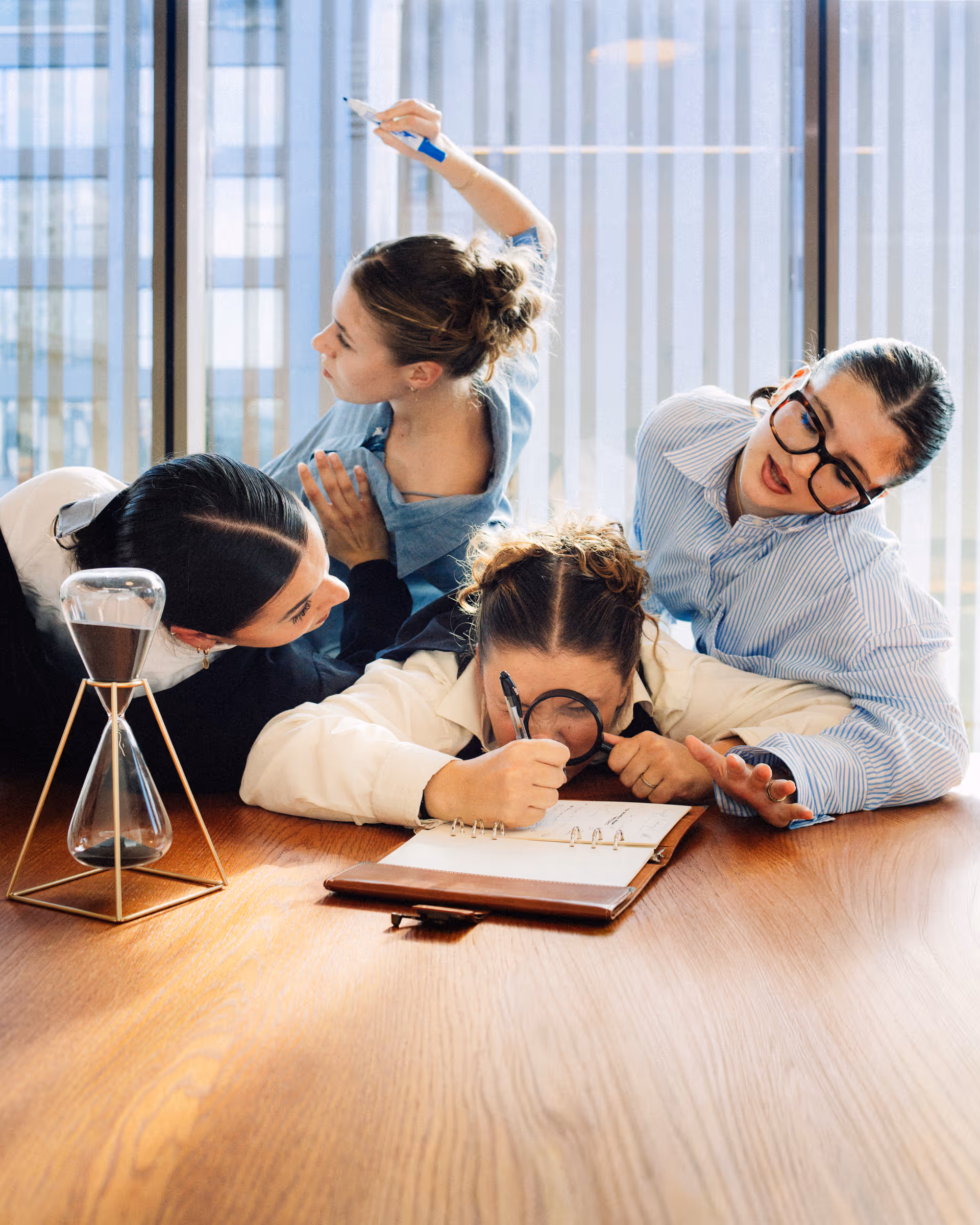 Four women collaborating intensely around a wooden table, one using a magnifying glass to examine a notebook closely.