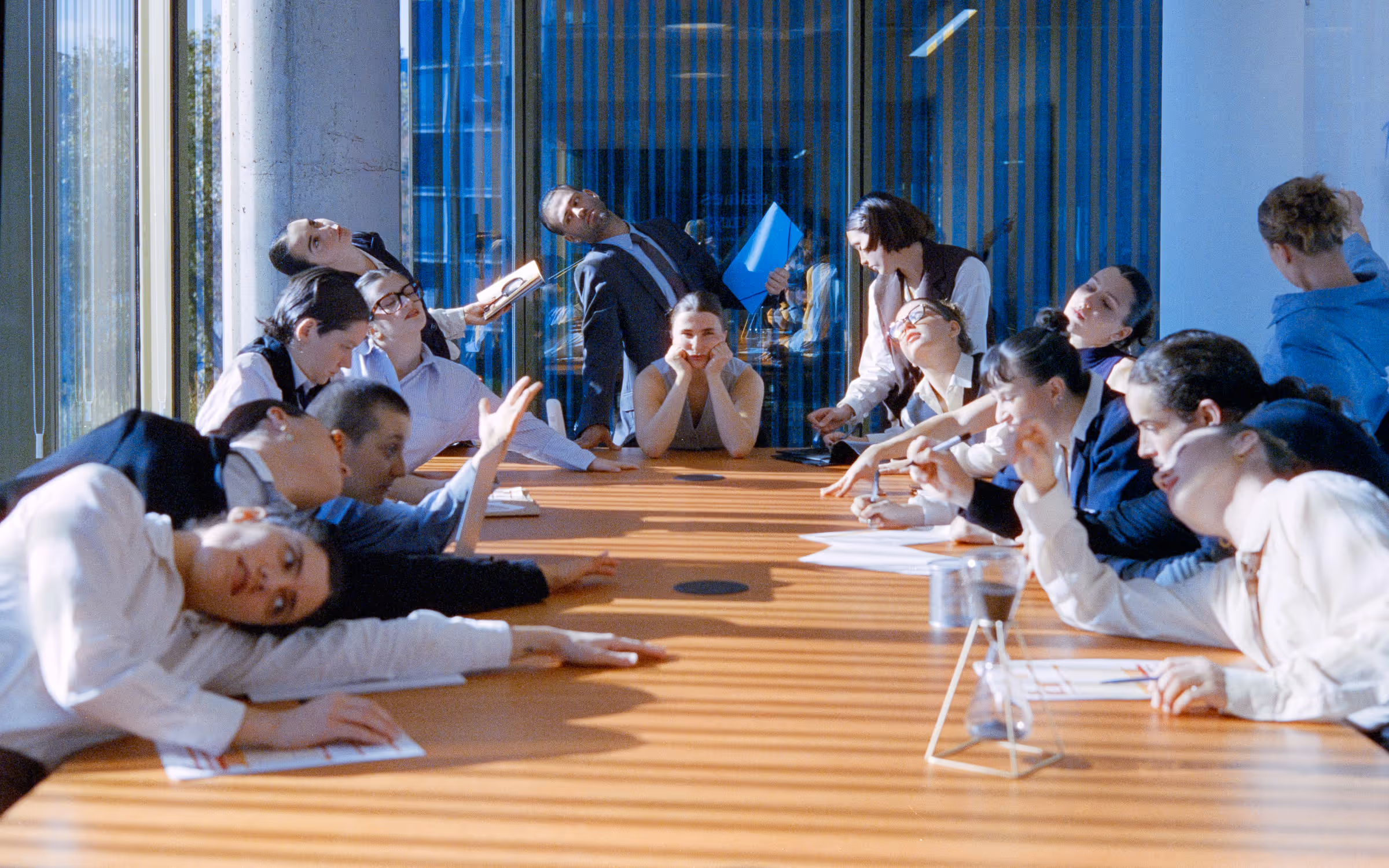 People around a conference table exhibiting signs of boredom and fatigue during a meeting in a sunlit office.