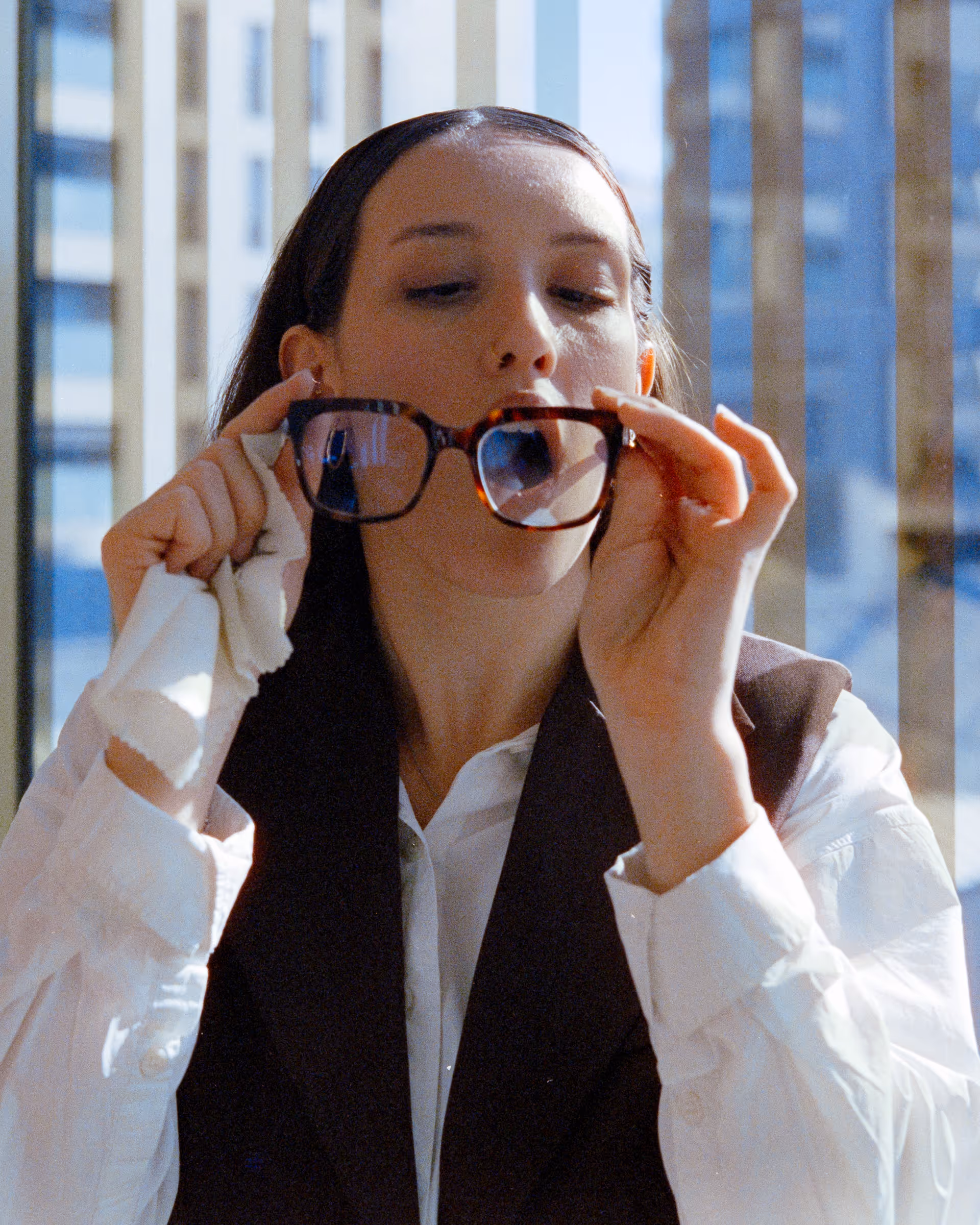 Woman holding and cleaning eyeglasses with a white cloth in front of a window with city buildings in the background.