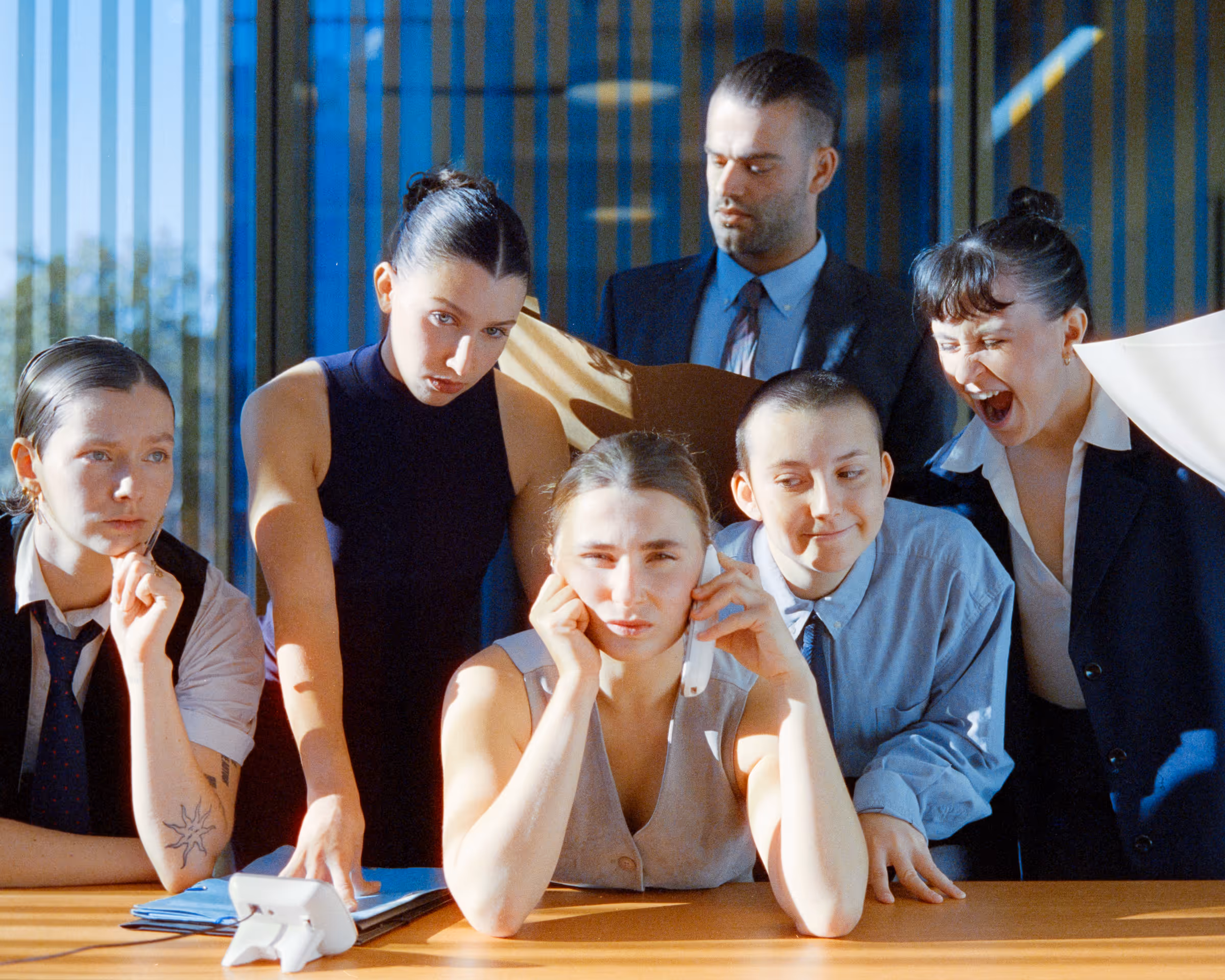 A group of five people in business attire encircle a woman on the phone looking stressed, while others appear concerned or shouting.