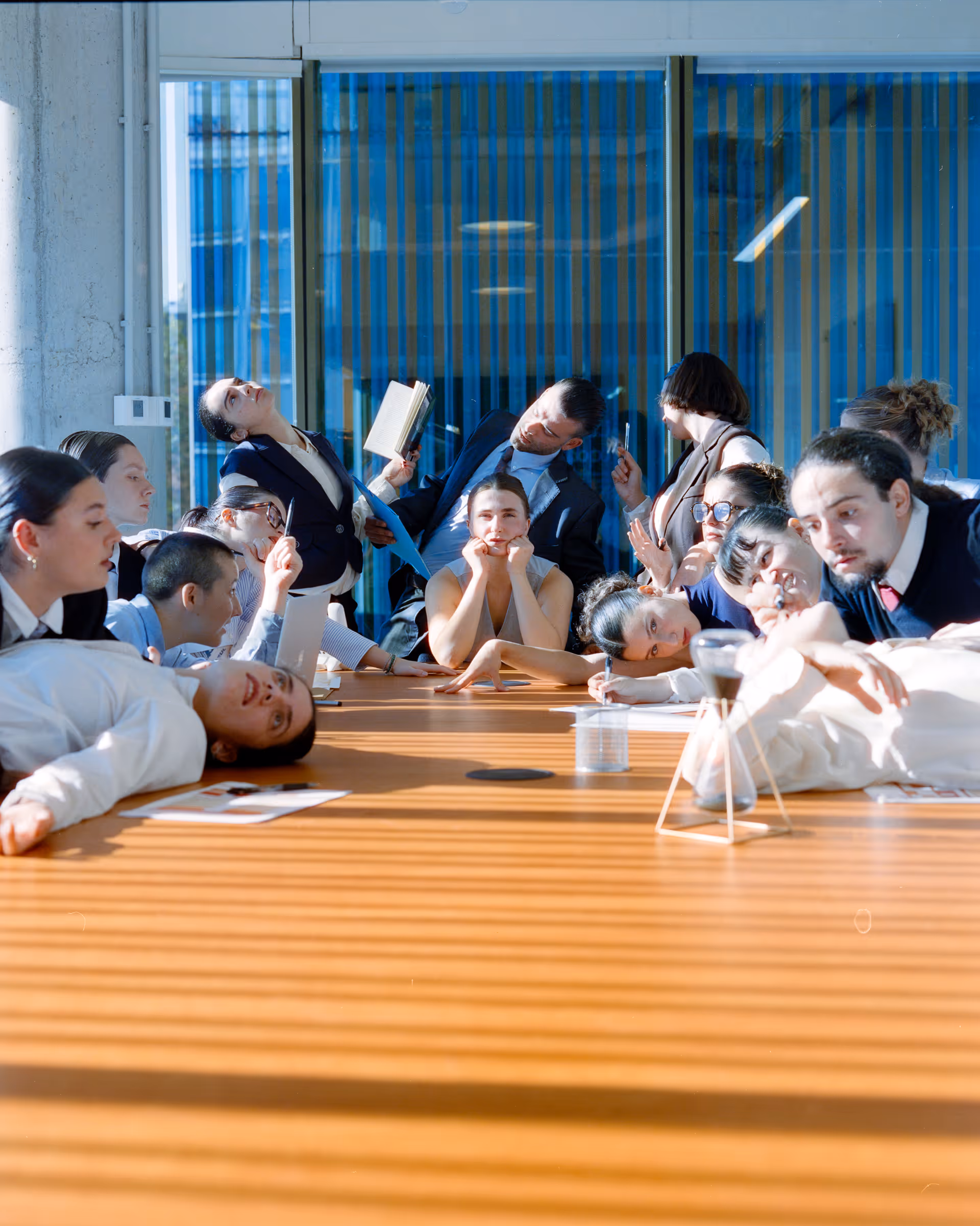 Group of diverse young adults in business attire gathered around a wooden conference table with notebooks, pens, and a sand timer, some appearing thoughtful and others engaged in animated discussion in a modern office setting.