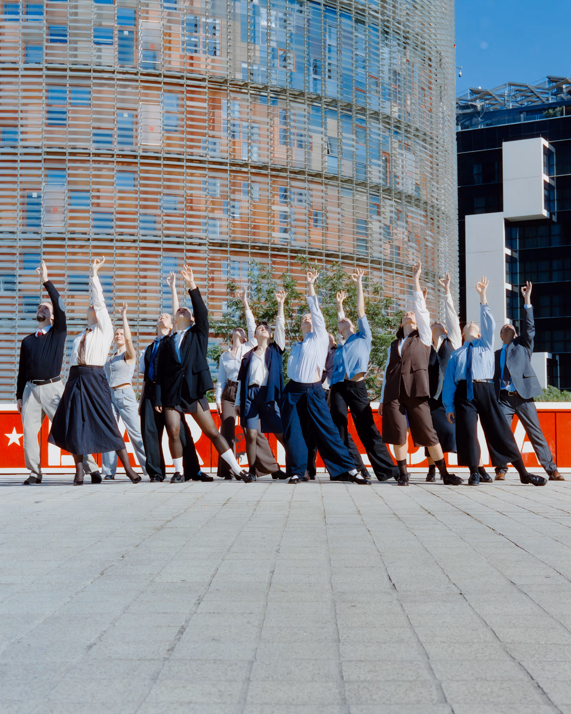 Group of people standing in a row outdoors with one arm raised upward, in front of modern glass buildings.