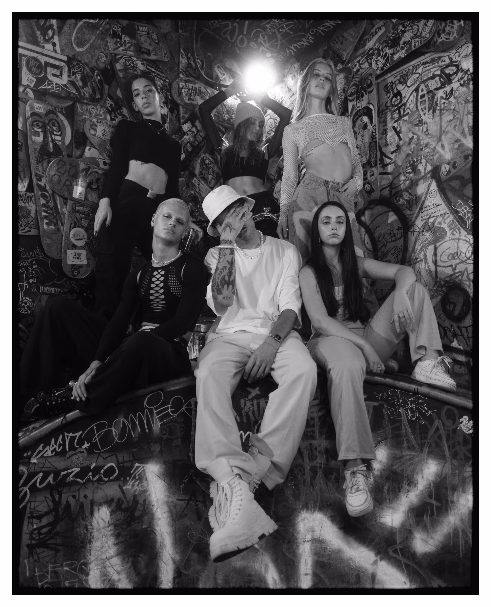 Black and white photo of six young adults posing in a graffiti-covered urban setting with edgy fashion styles and serious expressions.