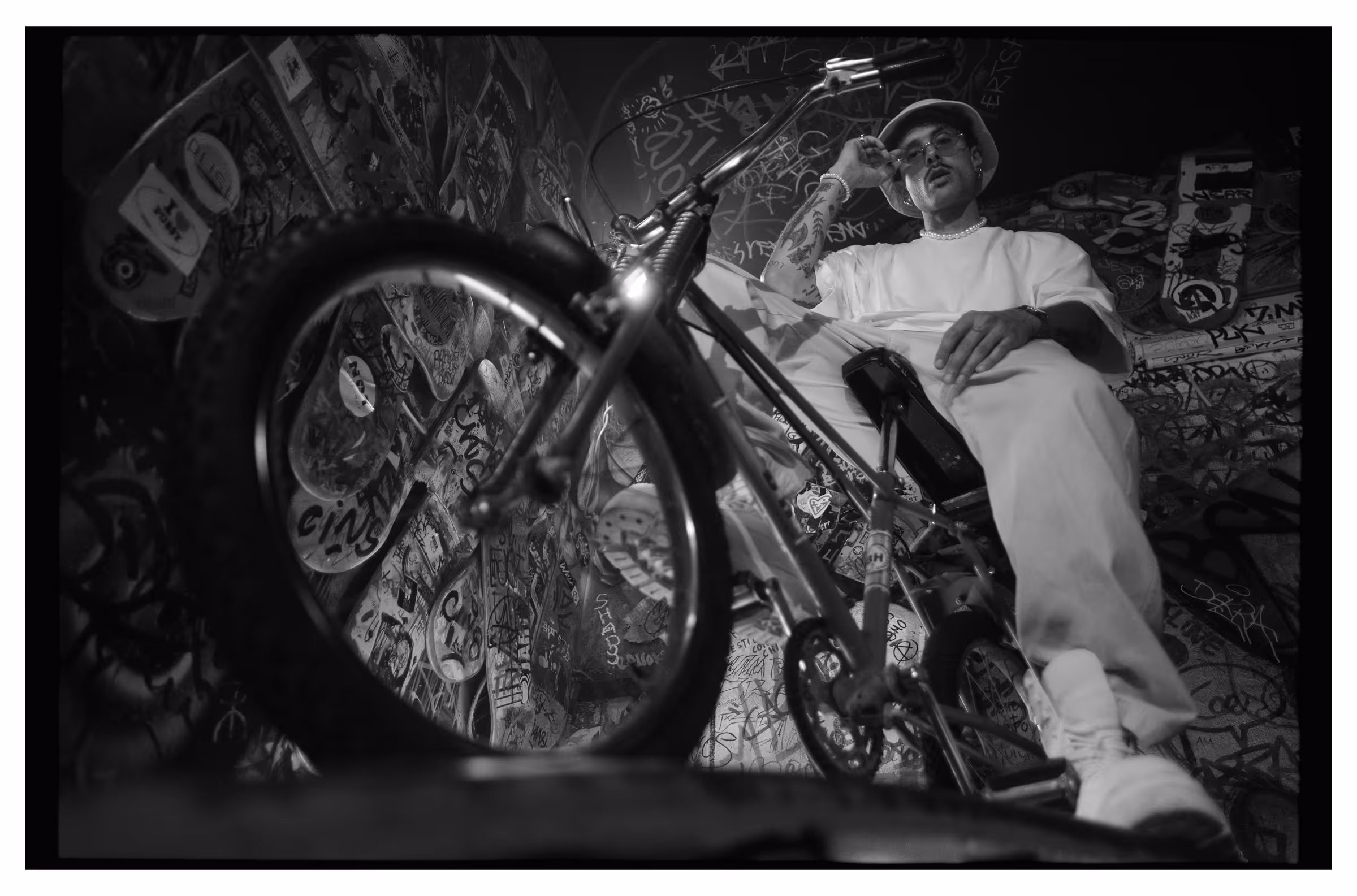 Man with tattoos and glasses sitting on a bicycle in a graffiti-covered room.