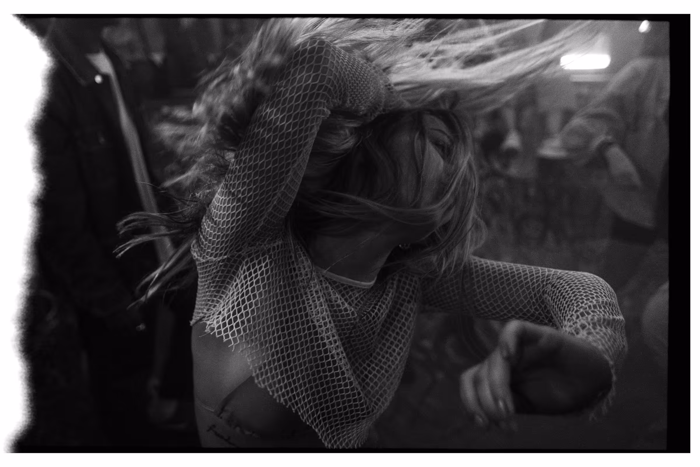 Black and white photo of a woman dancing energetically with hair flying and wearing a fishnet top in a dimly lit club.