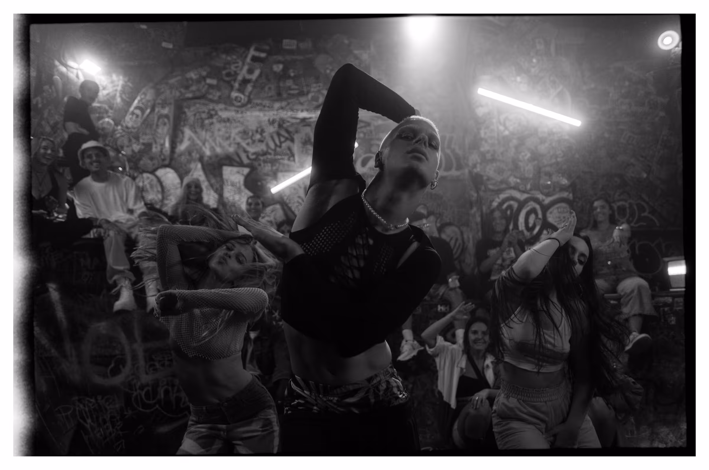 Black and white photo of three avant-garde dancers performing energetically in front of a graffiti-covered wall with an audience watching.