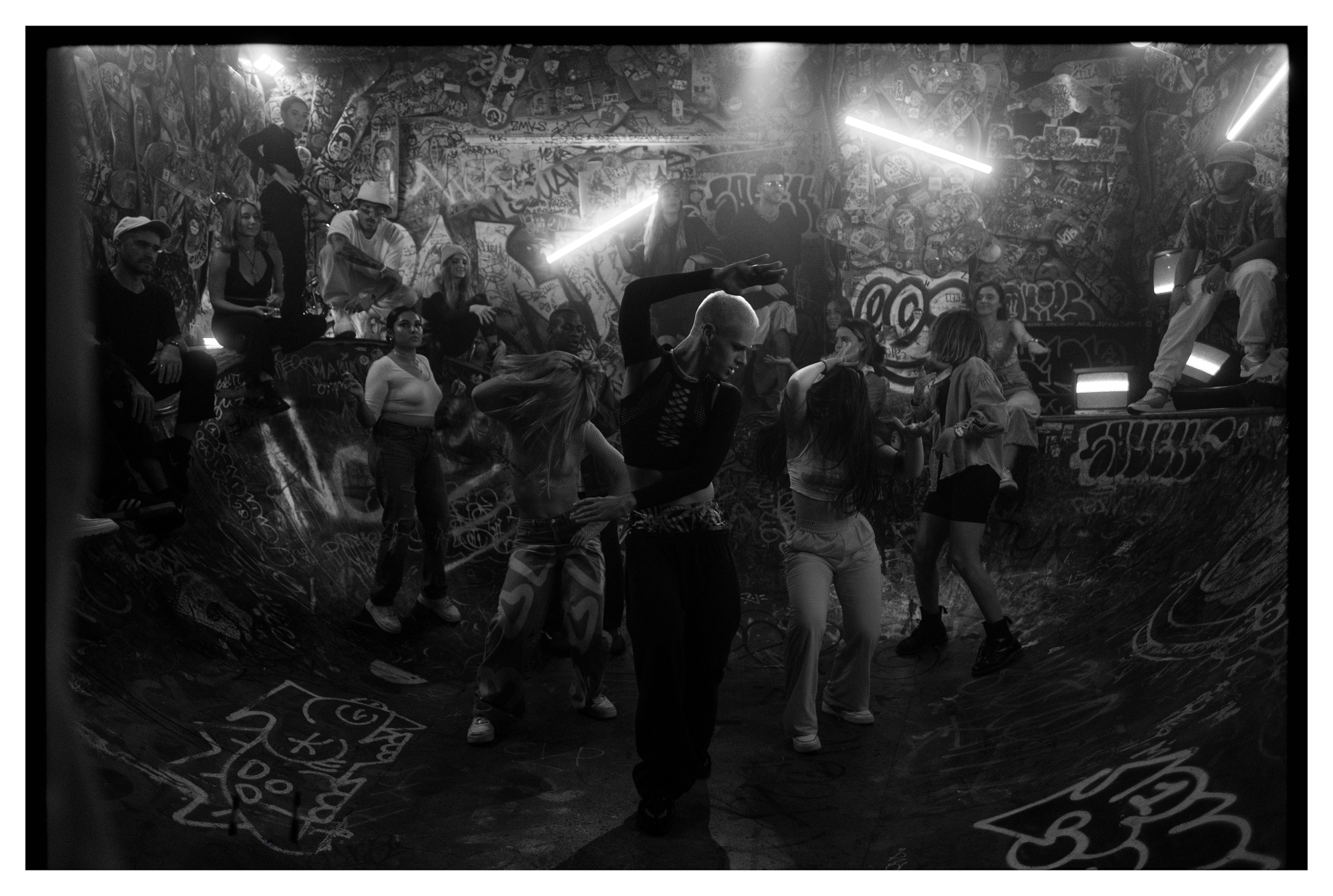 Black and white photo of a diverse group dancing and sitting in a graffiti-covered skatepark bowl with neon lights.