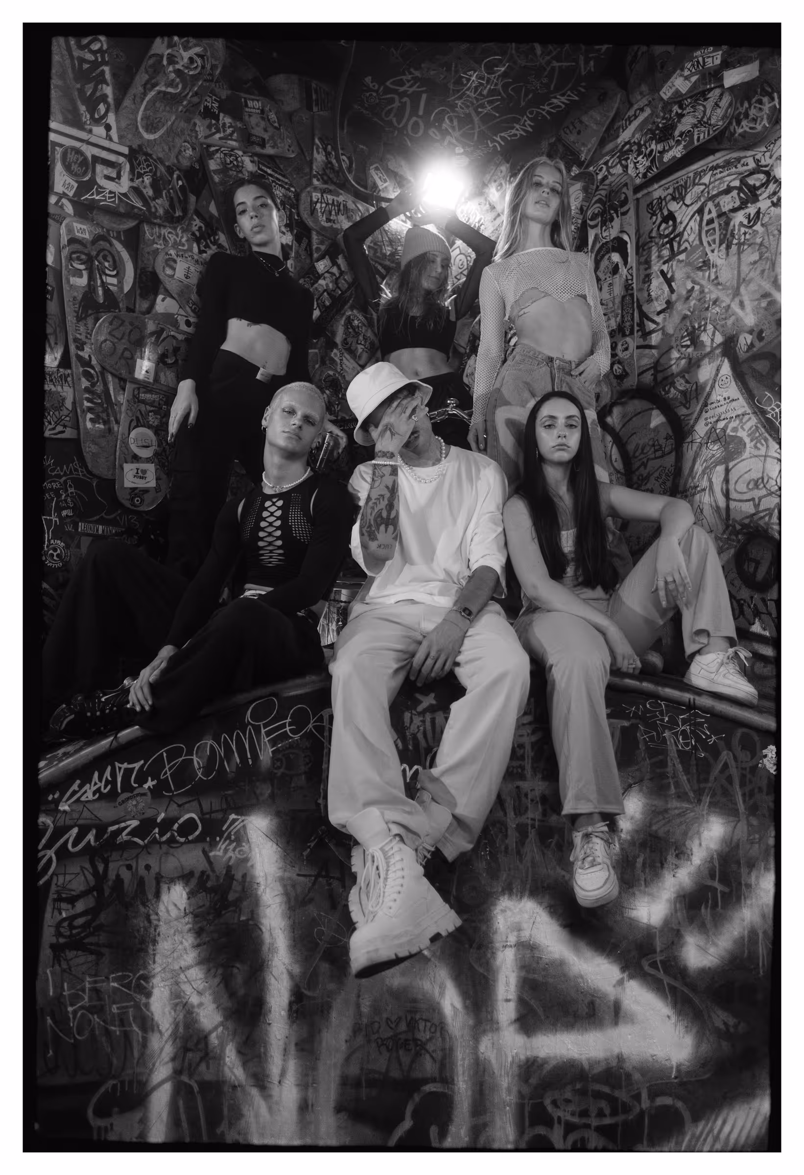 Black and white photo of six young people posing in a graffiti-covered stairwell, dressed in trendy streetwear with varied expressions.