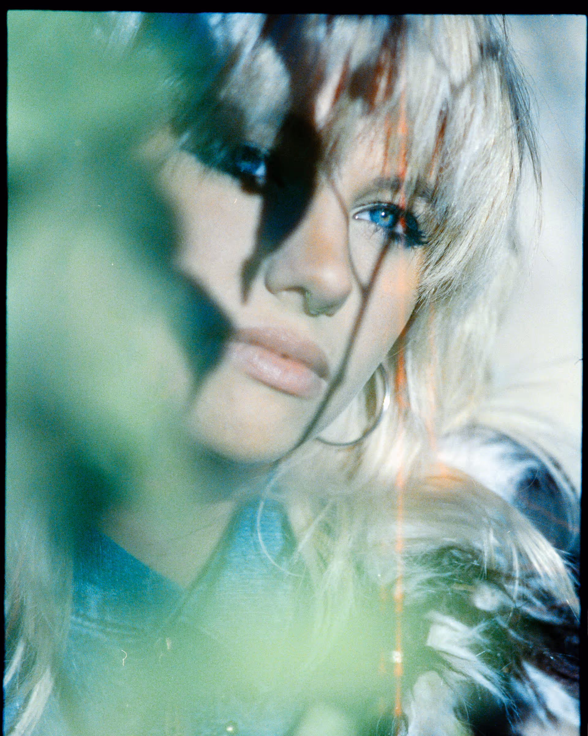 Close-up portrait of a blonde woman with blue eyes, partial green foliage in the foreground and light streaks creating a dreamy effect.