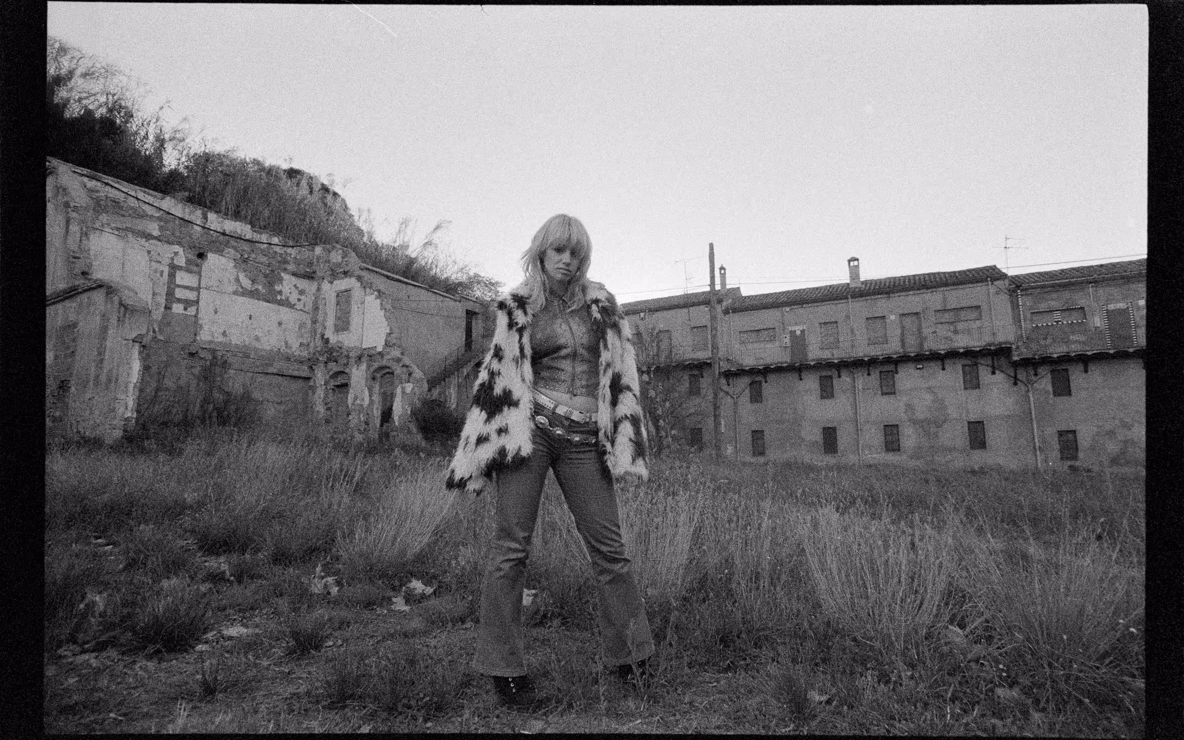 Black and white photo of a woman wearing a spotted fur coat and jeans standing in overgrown grass with dilapidated buildings in the background.
