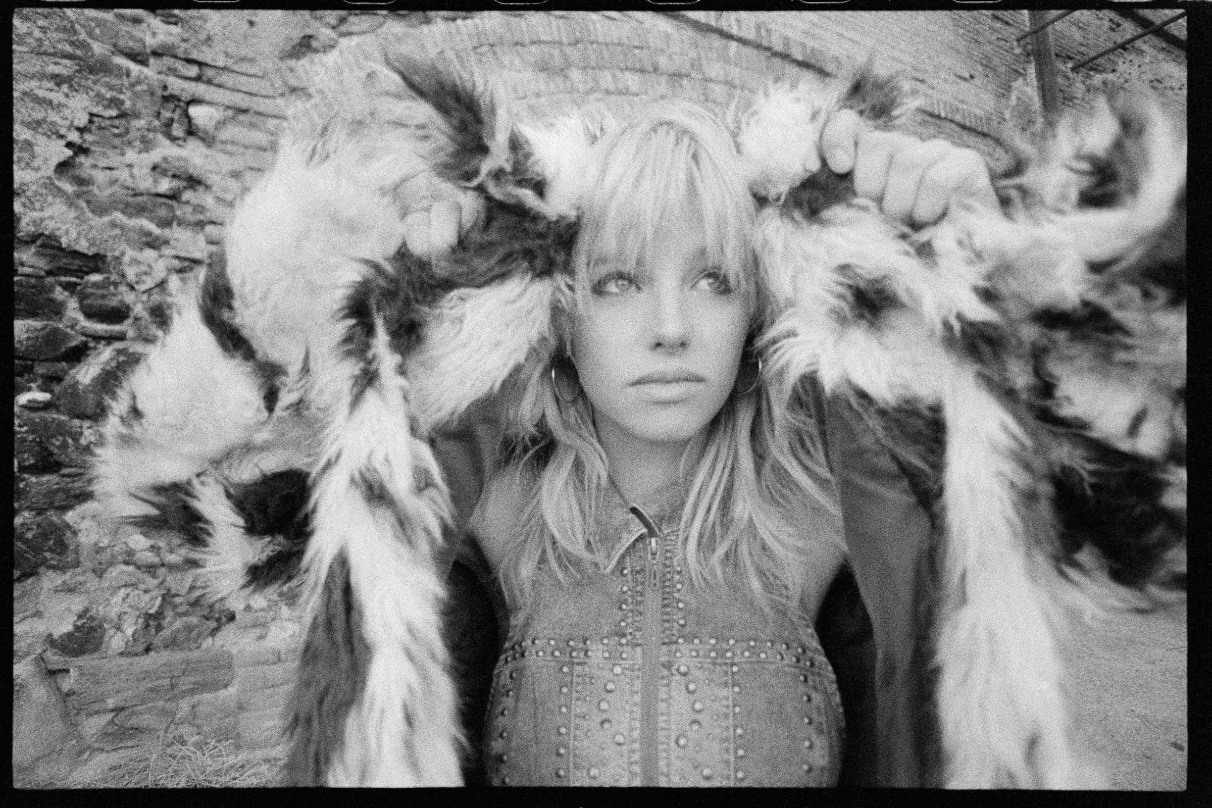 Young woman with long blonde hair holding a large furry coat above her head, standing in front of a stone wall.