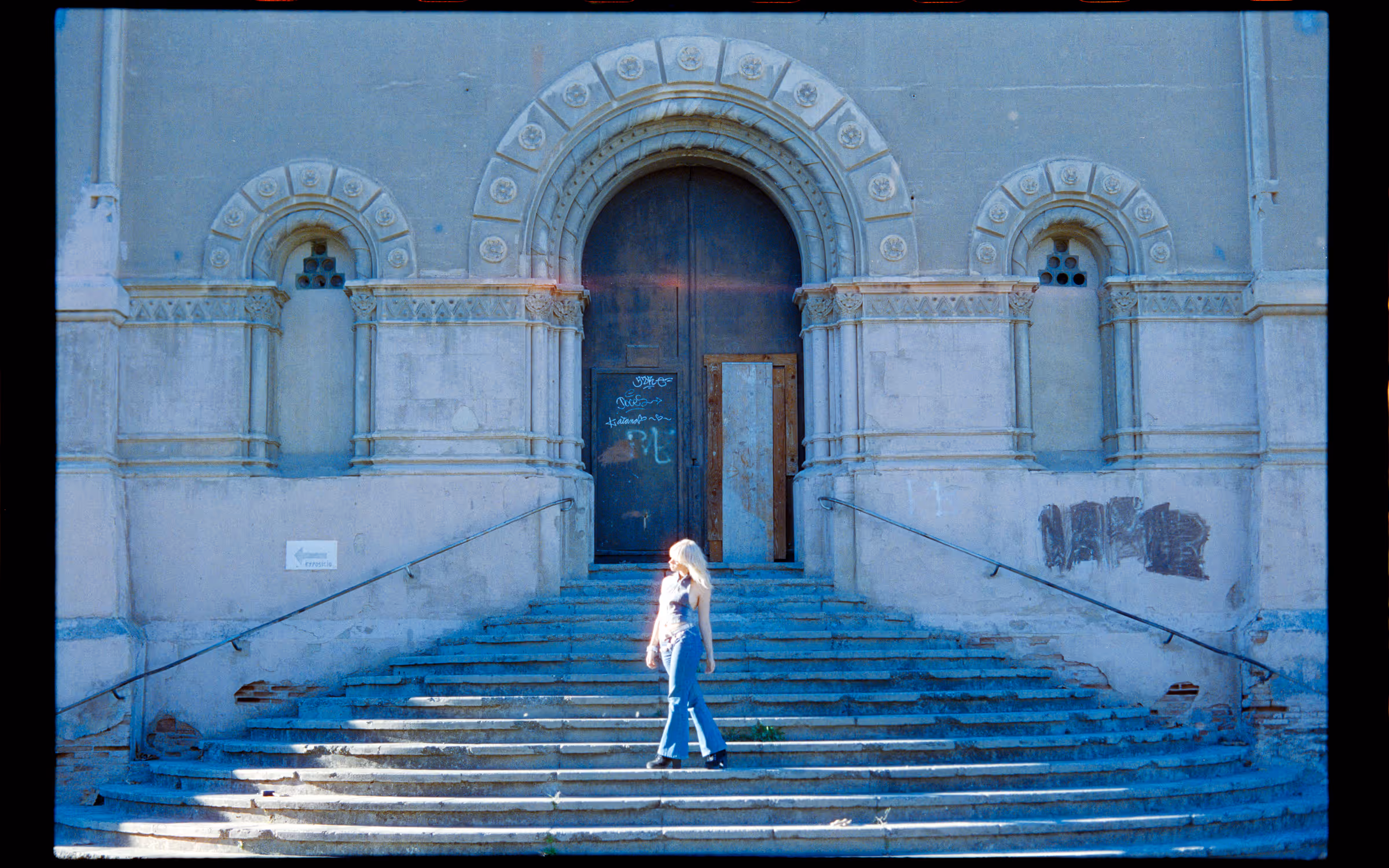 Woman with blonde hair walking down wide, worn stone steps in front of a large old building with ornate arched doorways and graffiti.