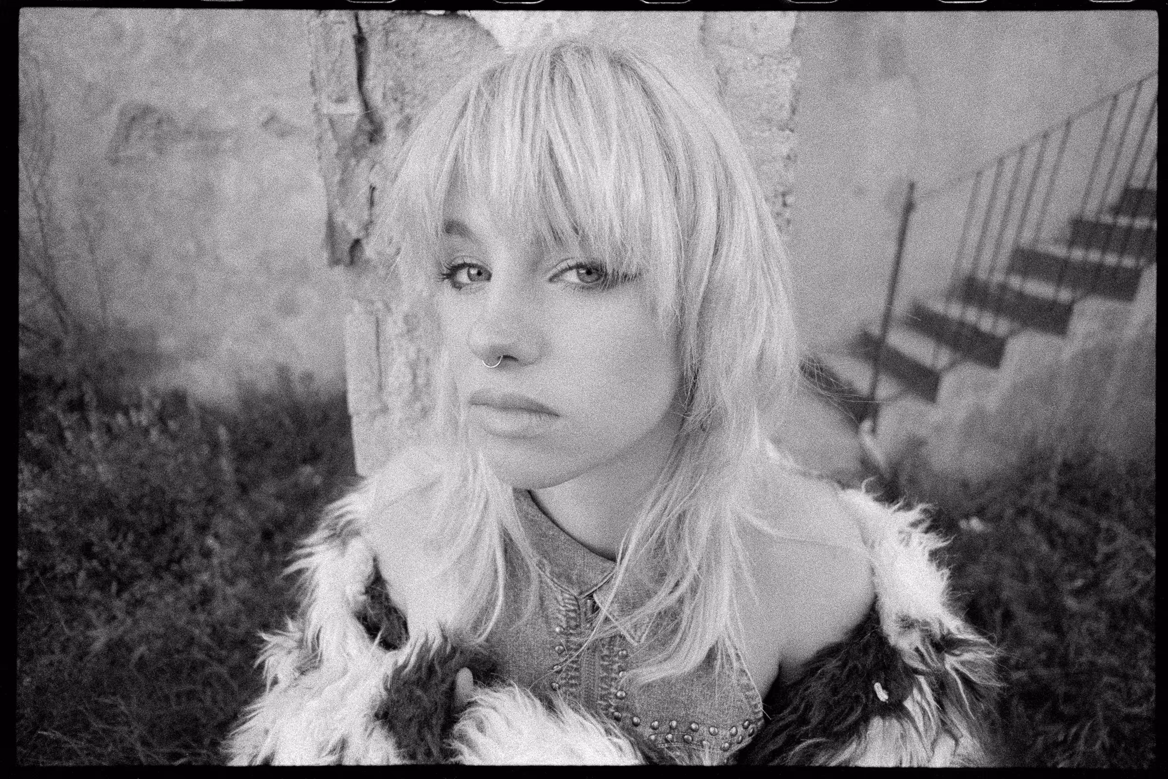Black and white close-up portrait of a young woman with light hair, a nose ring, and a fur-like jacket, standing outdoors near a stone wall and stairs.