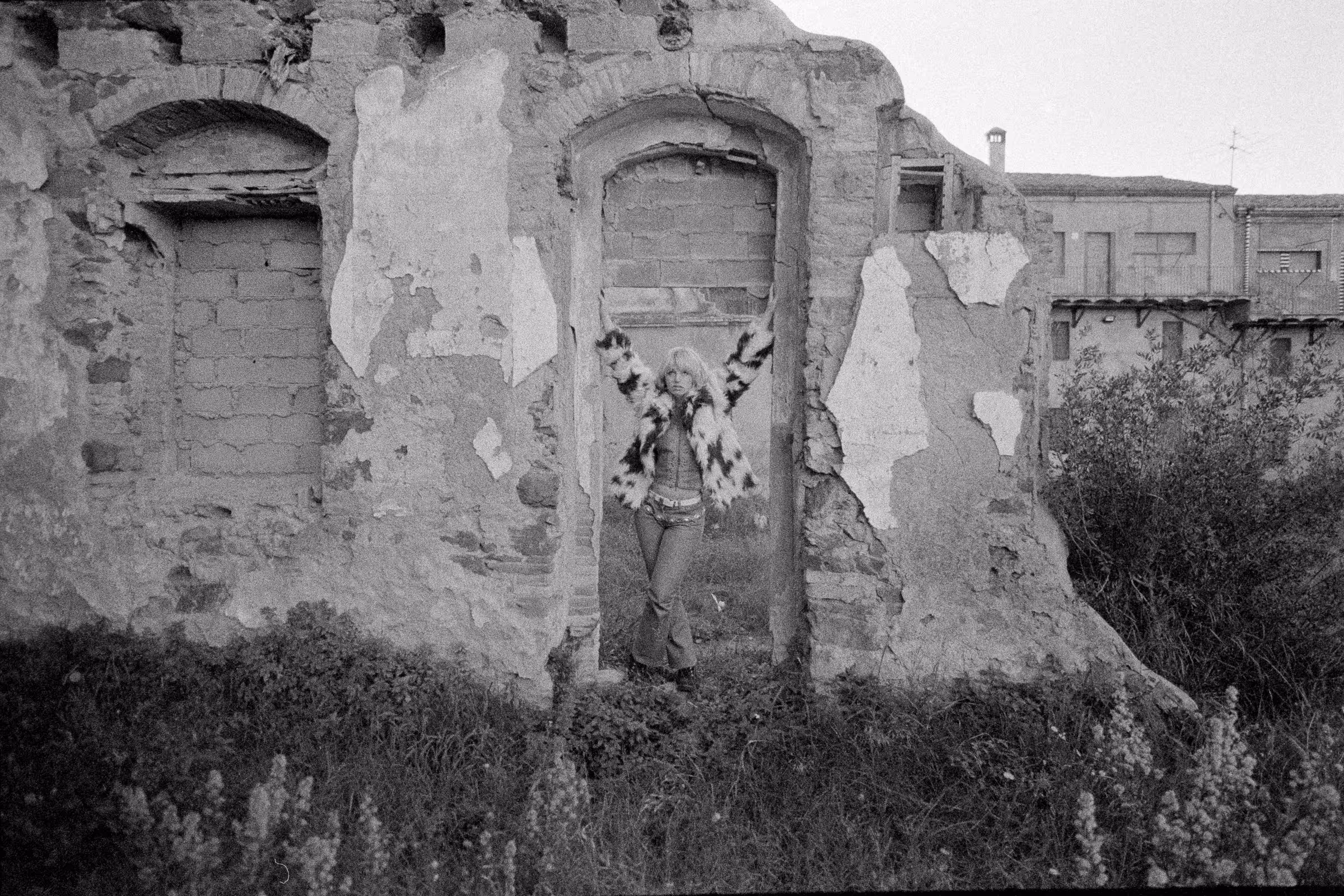 Person wearing a patterned fur jacket standing with arms raised in the doorway of a crumbling stone wall.
