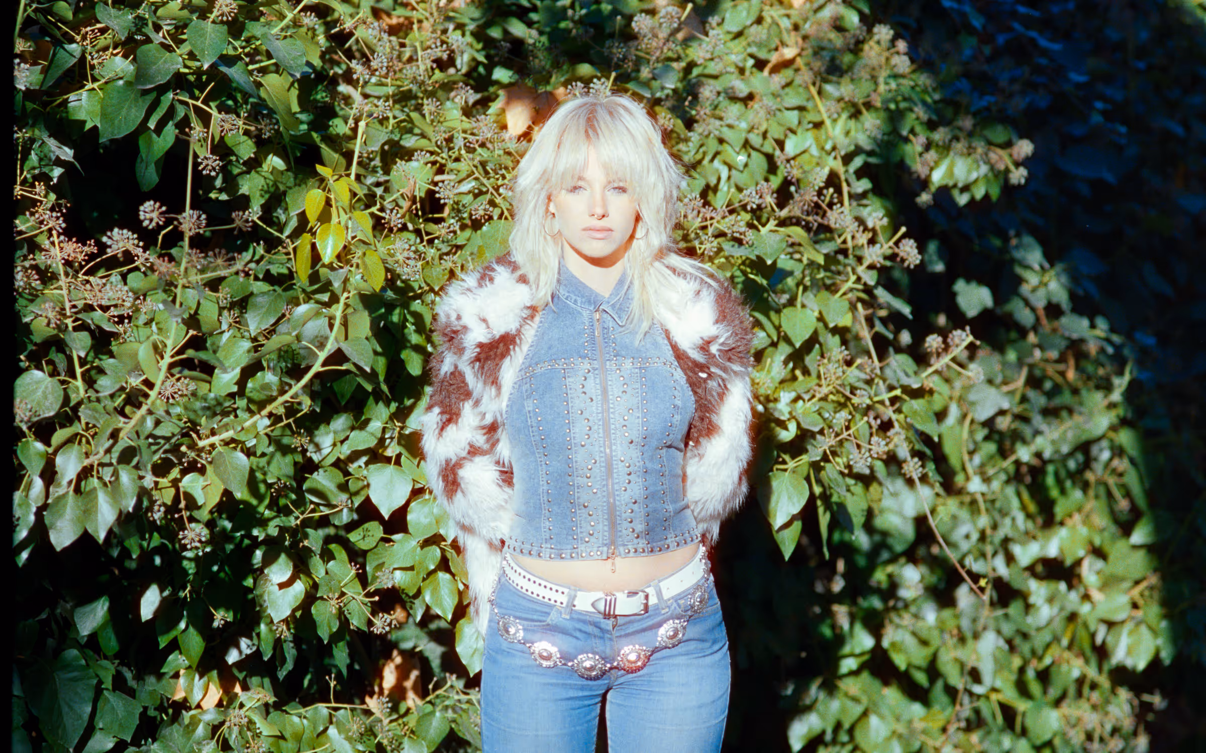 Blonde woman wearing a studded denim jacket with a white and brown fur coat and decorative belt, standing in front of green foliage.