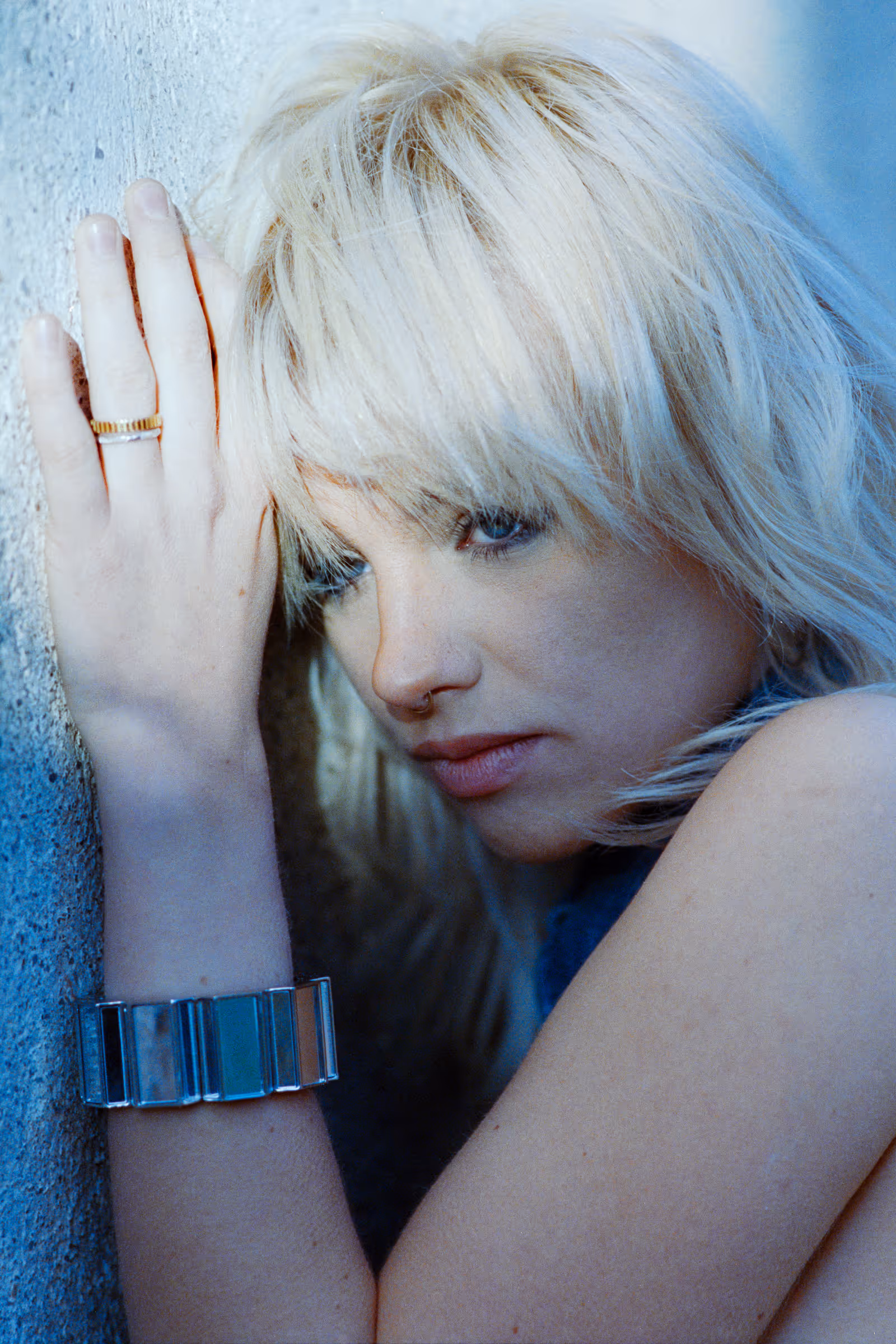 Close-up of a blonde woman with a nose ring and bracelet leaning her head against a textured wall.