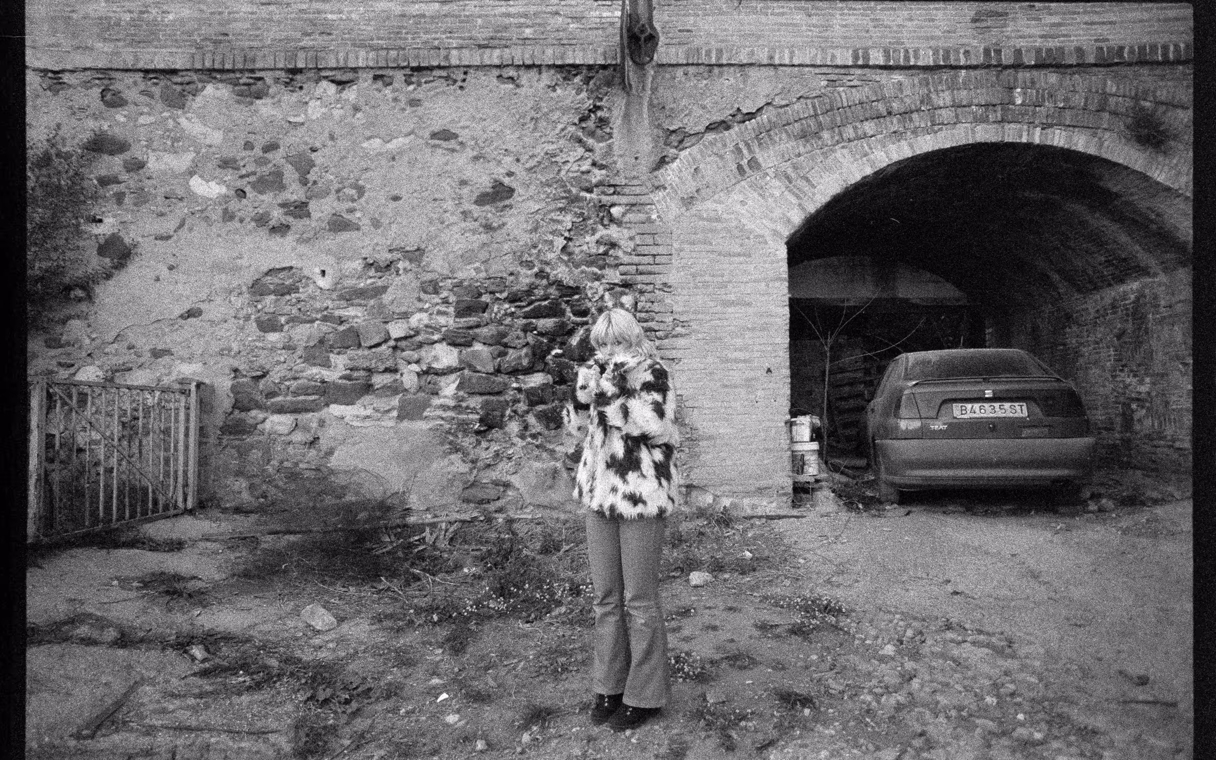 Person wearing a spotted fur coat and pants stands in front of a rough stone wall and an archway with a parked car inside.