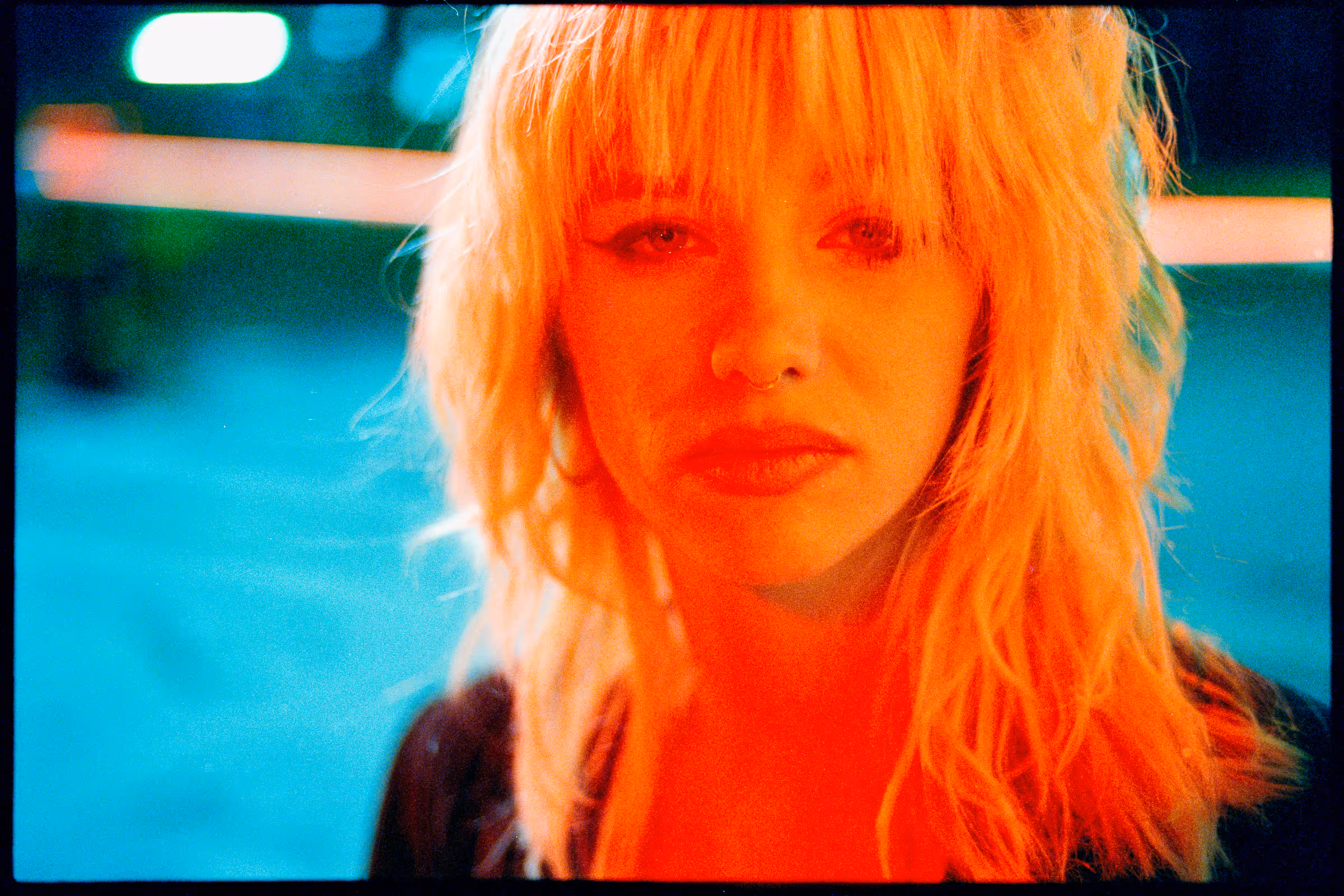 Portrait of a woman with blonde hair and a nose ring illuminated by orange and blue lighting.