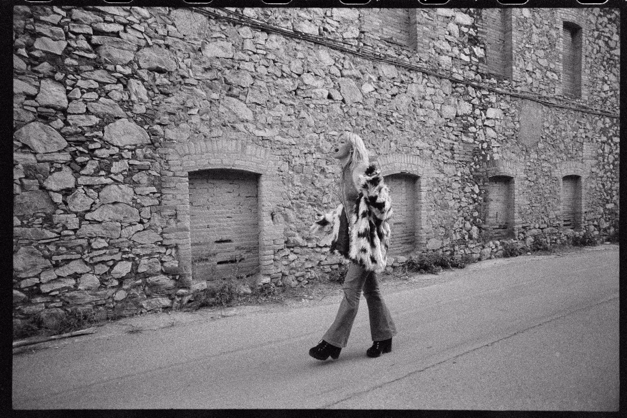 Black and white photo of a person wearing a patterned fur coat and platform shoes walking along a stone wall with barricaded window openings.