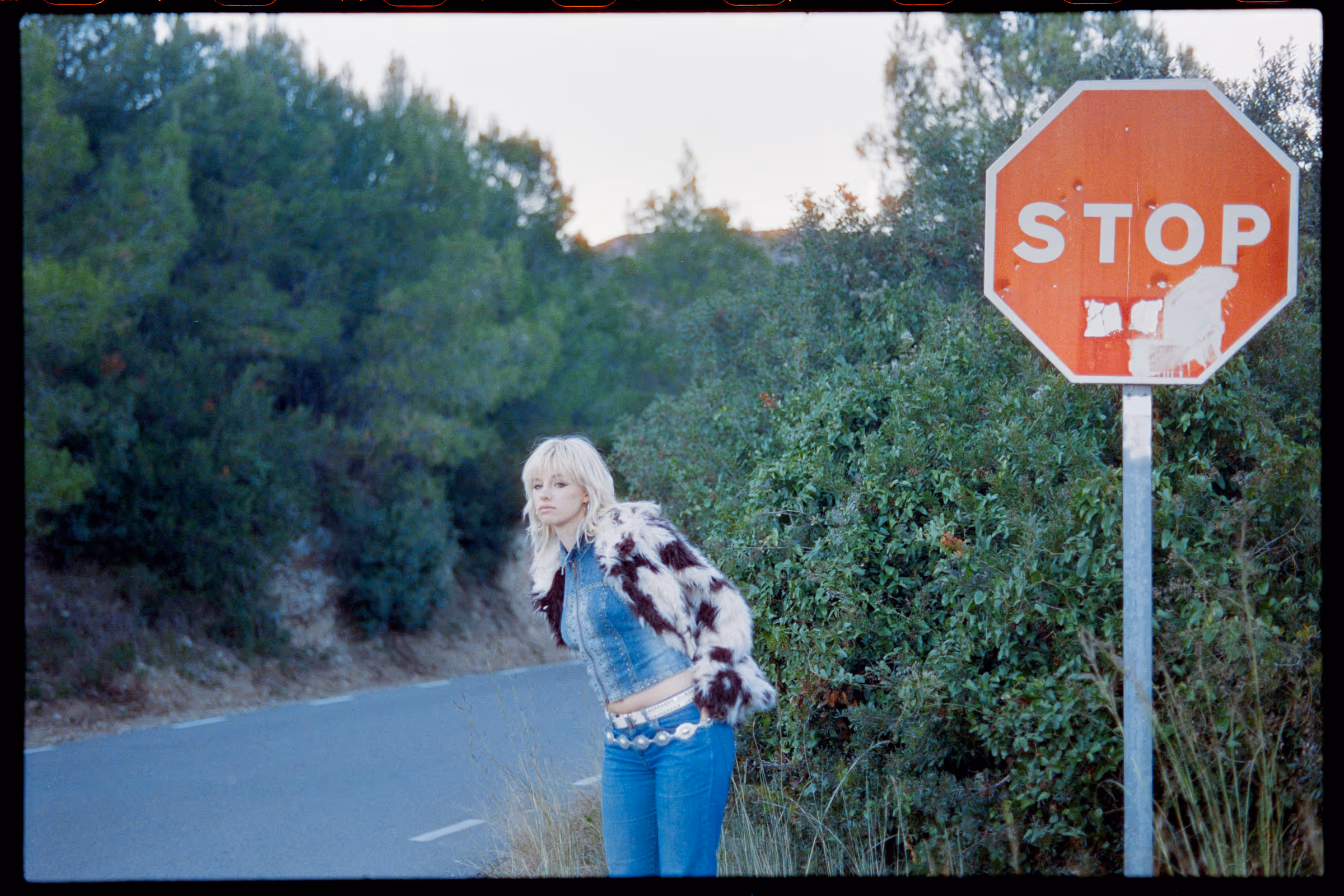 Blonde woman in a fur jacket and denim jeans standing near a worn stop sign beside a road with dense green bushes.