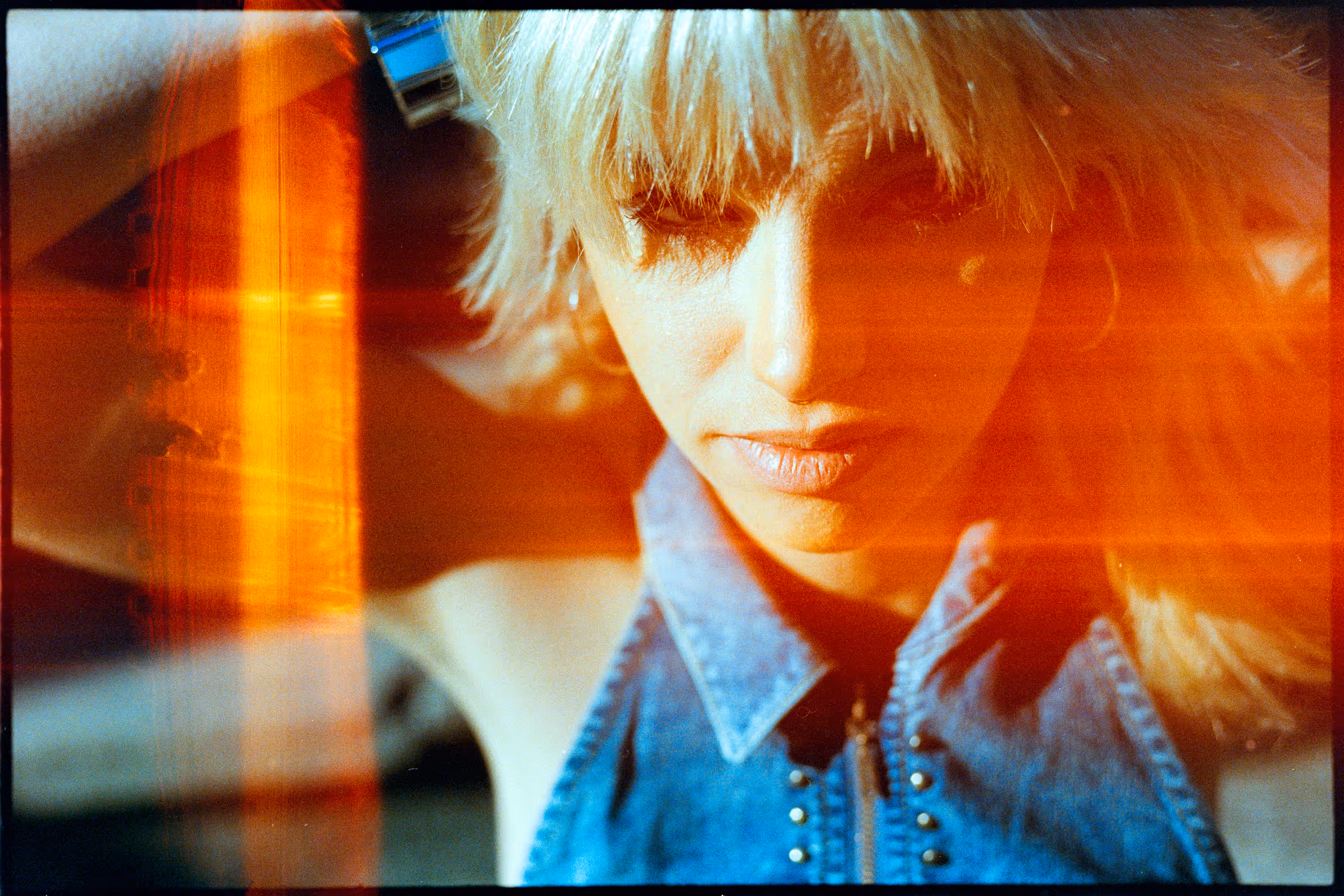 Close-up of a woman with blonde hair and bangs wearing a blue denim sleeveless top, with orange light streaks across the image.