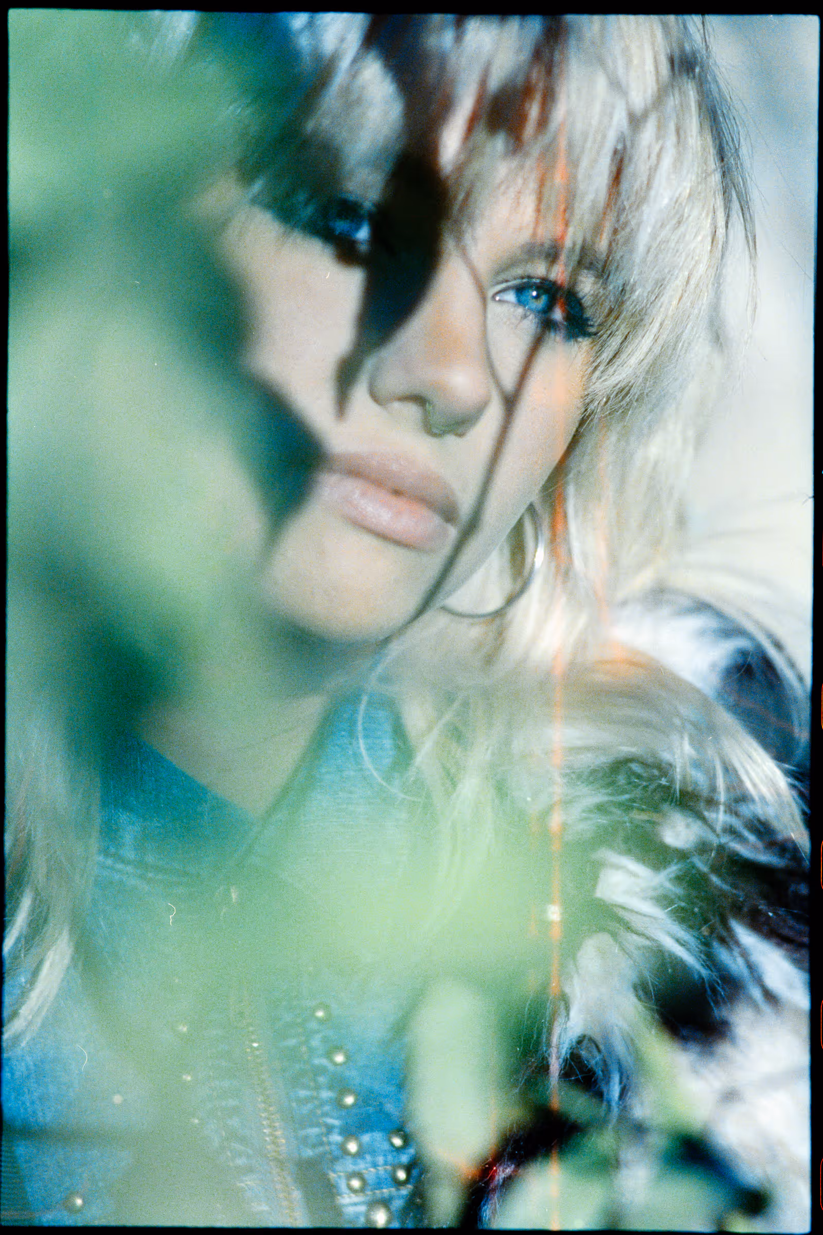 Close-up portrait of a woman with blonde hair, blue eyes, and hoop earrings, partially obscured by green foliage.