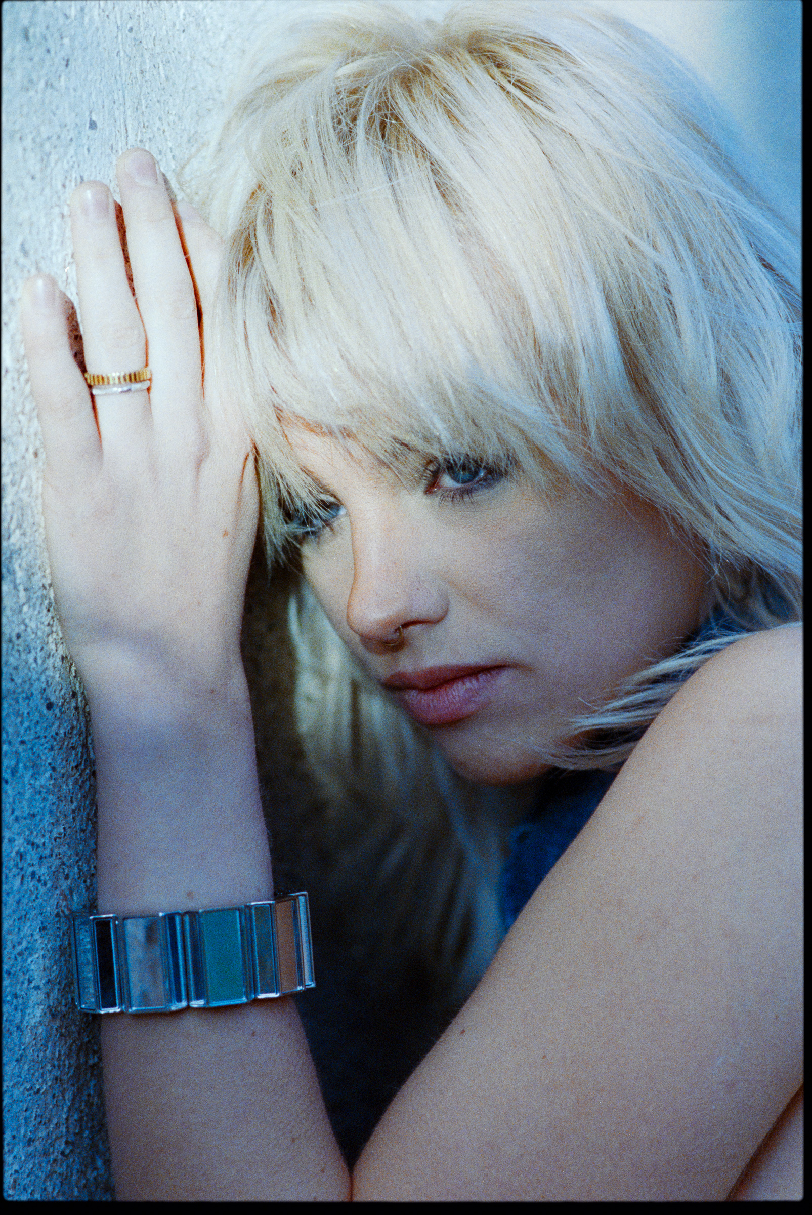 Close-up of a woman with platinum blonde hair resting her head against a textured wall, wearing a silver bracelet and rings.