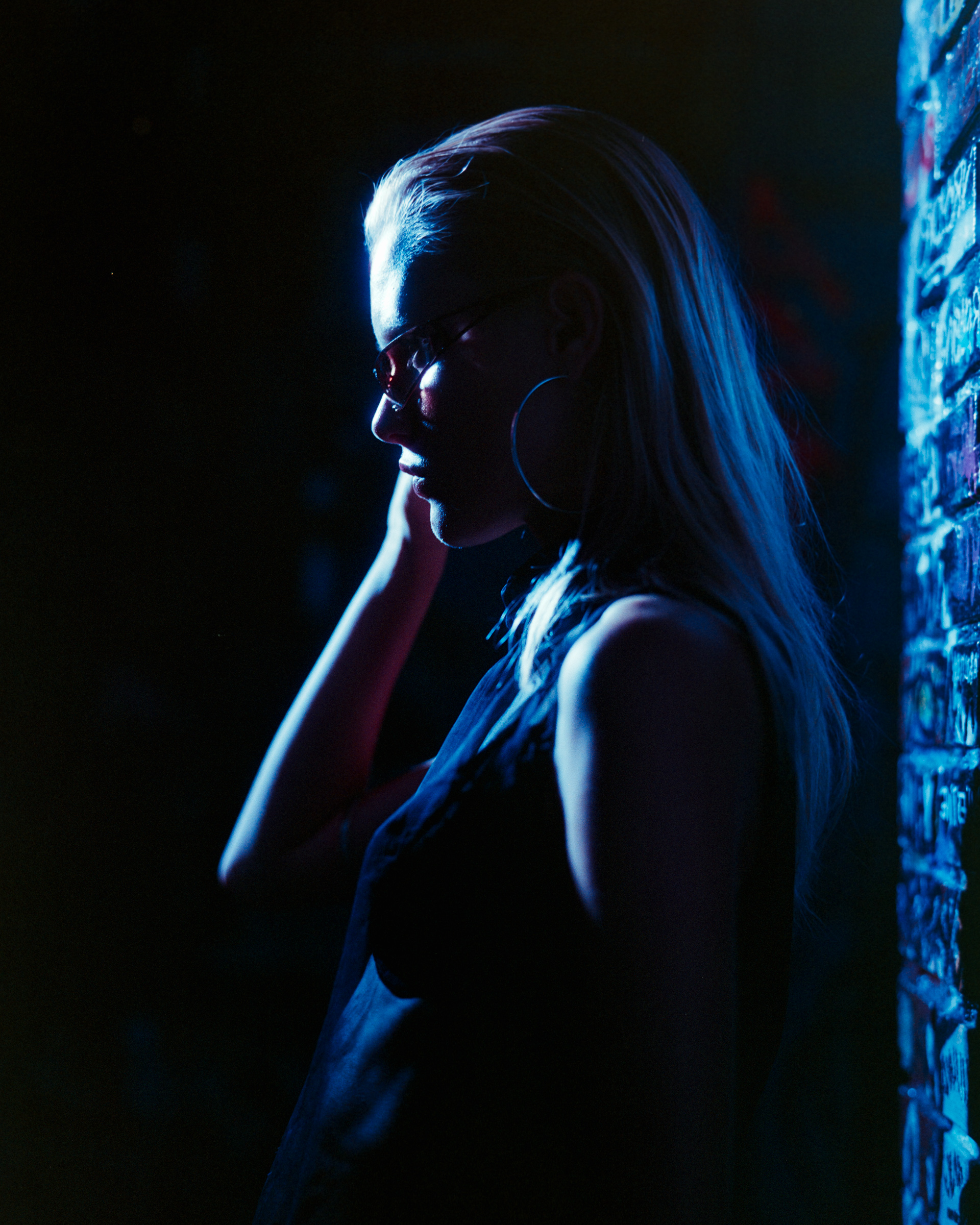 Profile of a woman with long hair and large hoop earrings, illuminated by blue light against a dark background and a graffiti-covered wall.