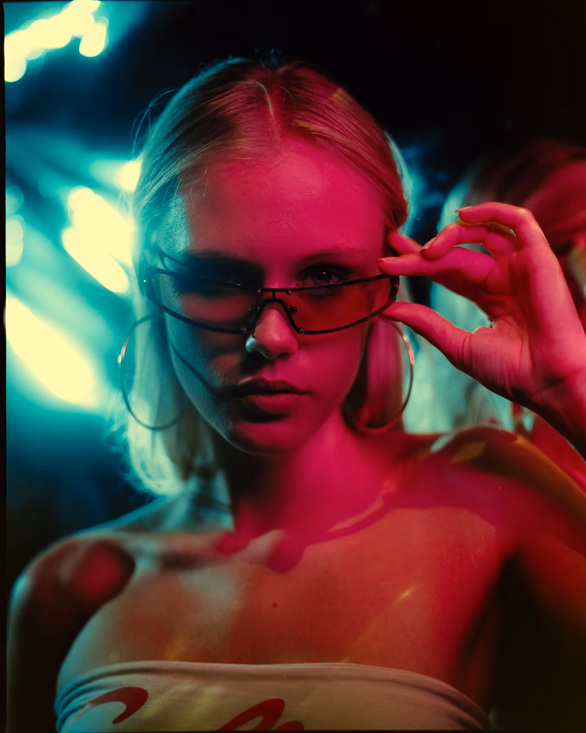 Young woman with blonde hair wearing hoop earrings and partially lowering red-tinted sunglasses under vibrant pink and blue lighting.