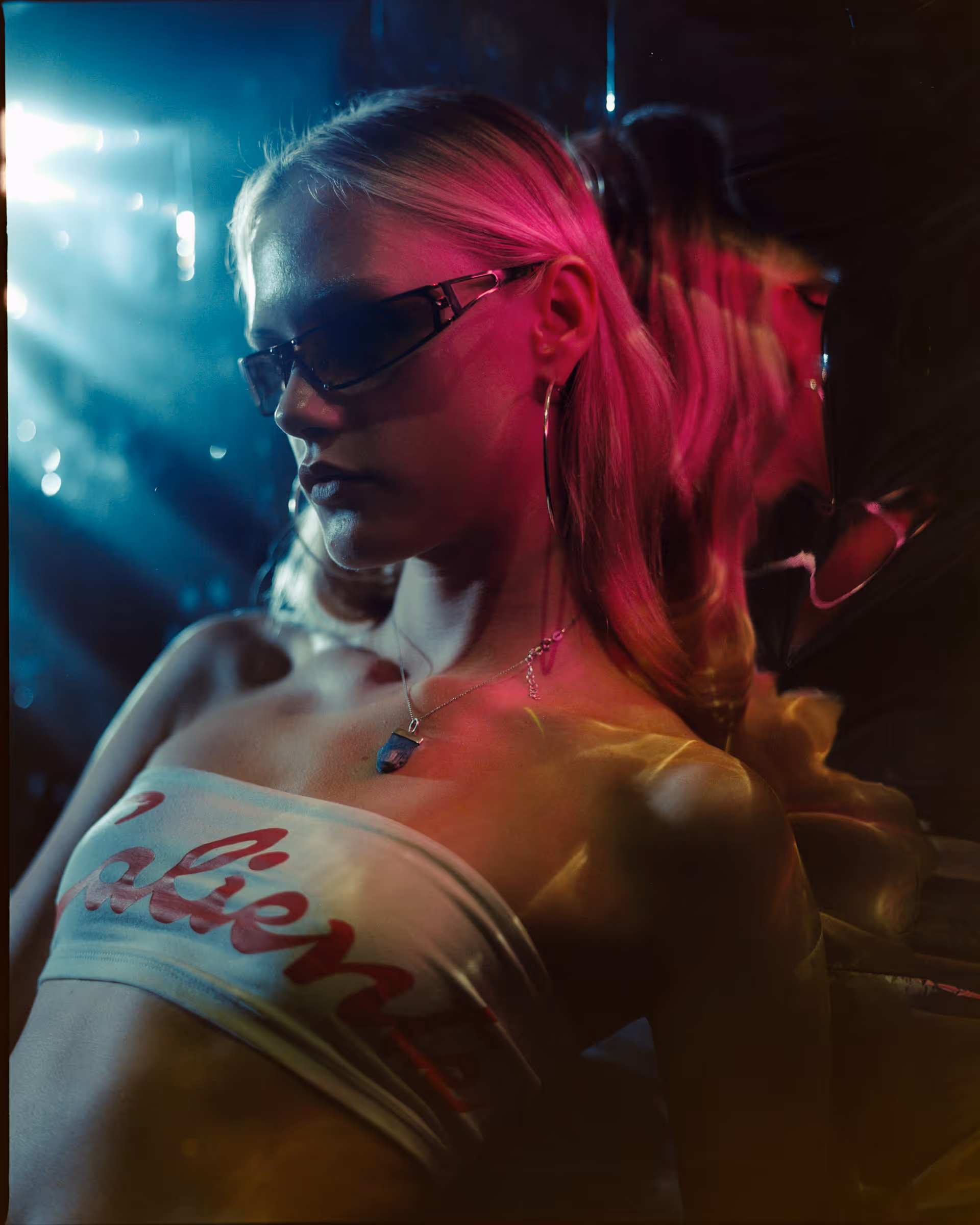 Woman with blonde hair wearing sunglasses, hoop earrings, and a white tube top with red text, illuminated by pink and blue lighting.