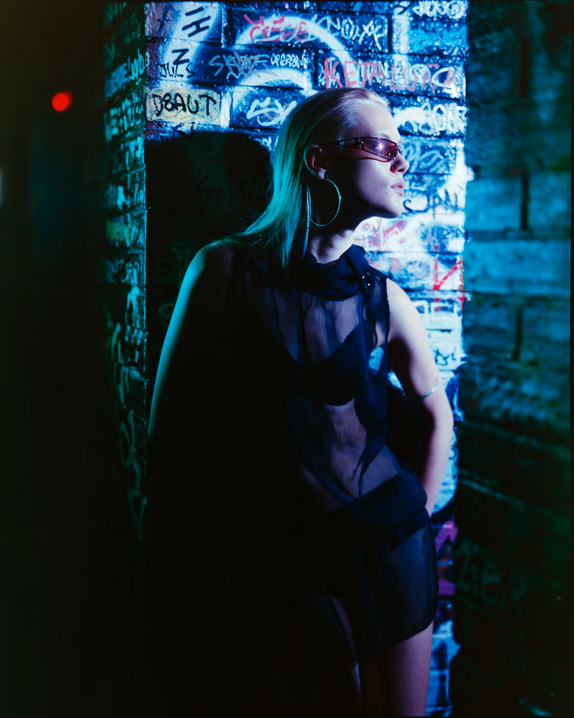 Woman with slicked-back hair wearing red sunglasses, large hoop earrings, and a sheer black outfit leaning against a graffiti-covered wall in blue lighting.