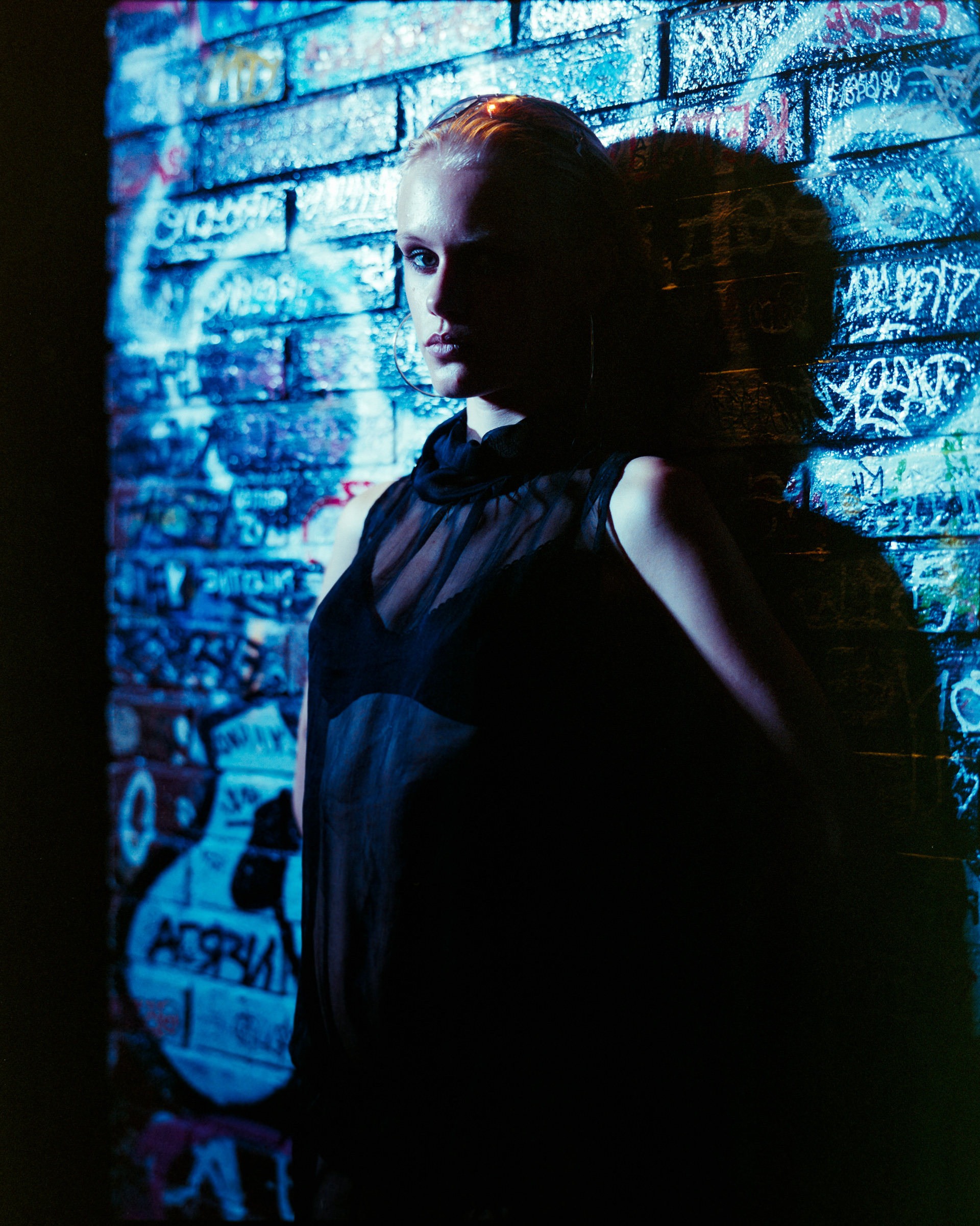 Person with slicked-back hair wearing a sheer black top, standing against a graffiti-covered brick wall illuminated by blue light.