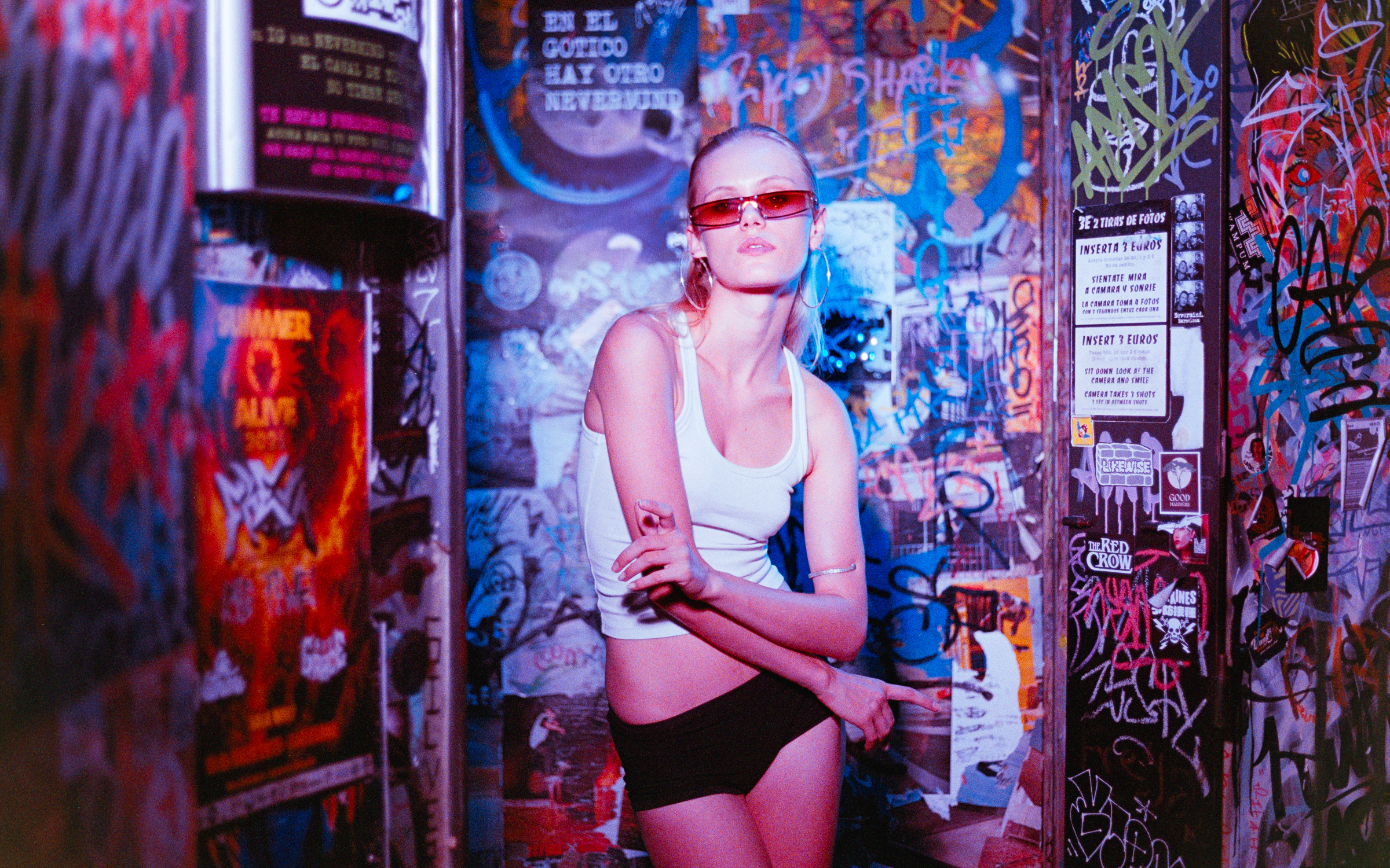 Young woman with red tinted sunglasses and hoop earrings posing with folded arms in a graffiti-covered room.