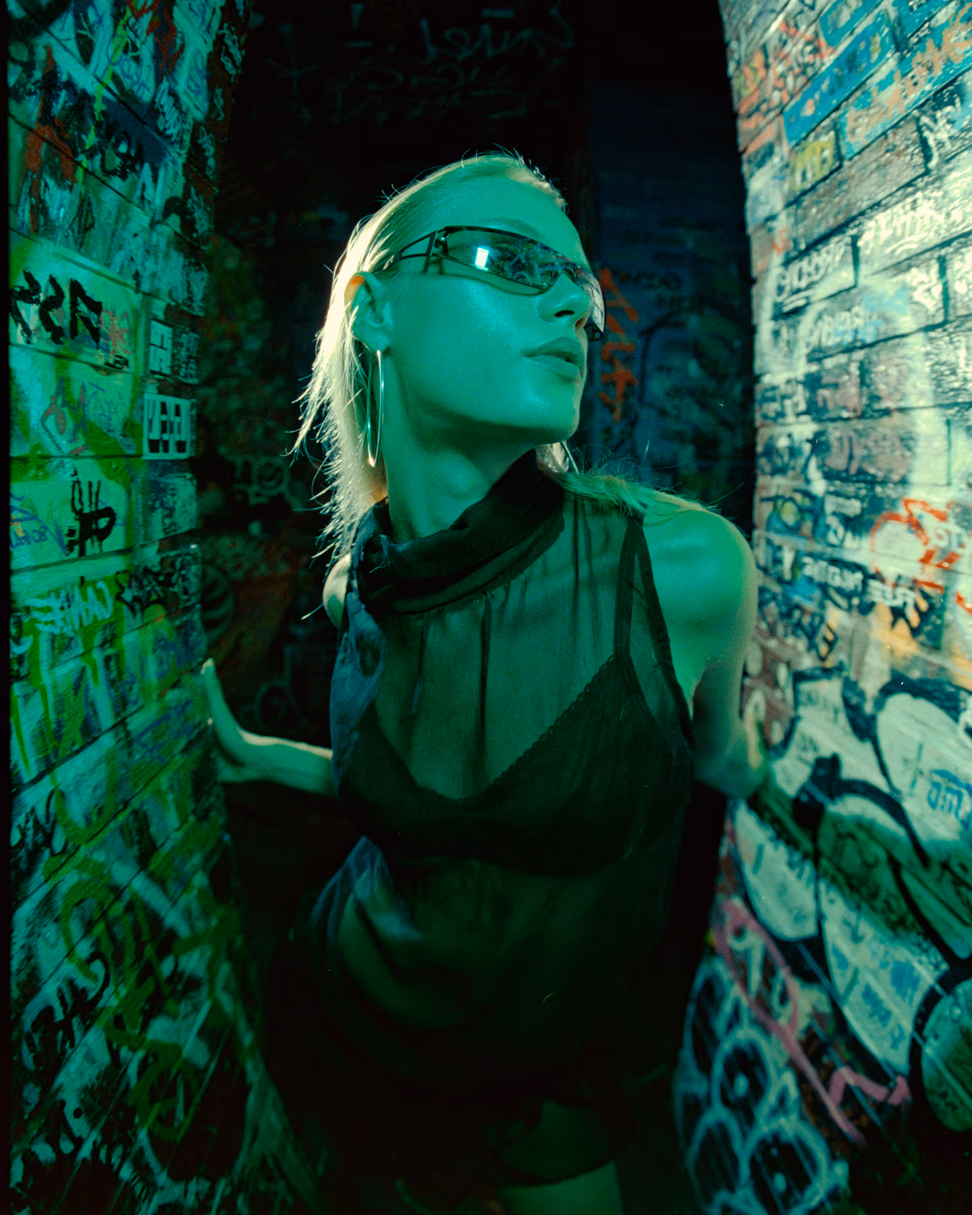 Young woman with blonde hair and large hoop earrings wearing translucent black clothing and sunglasses, posing between two graffiti-covered brick walls under green lighting.