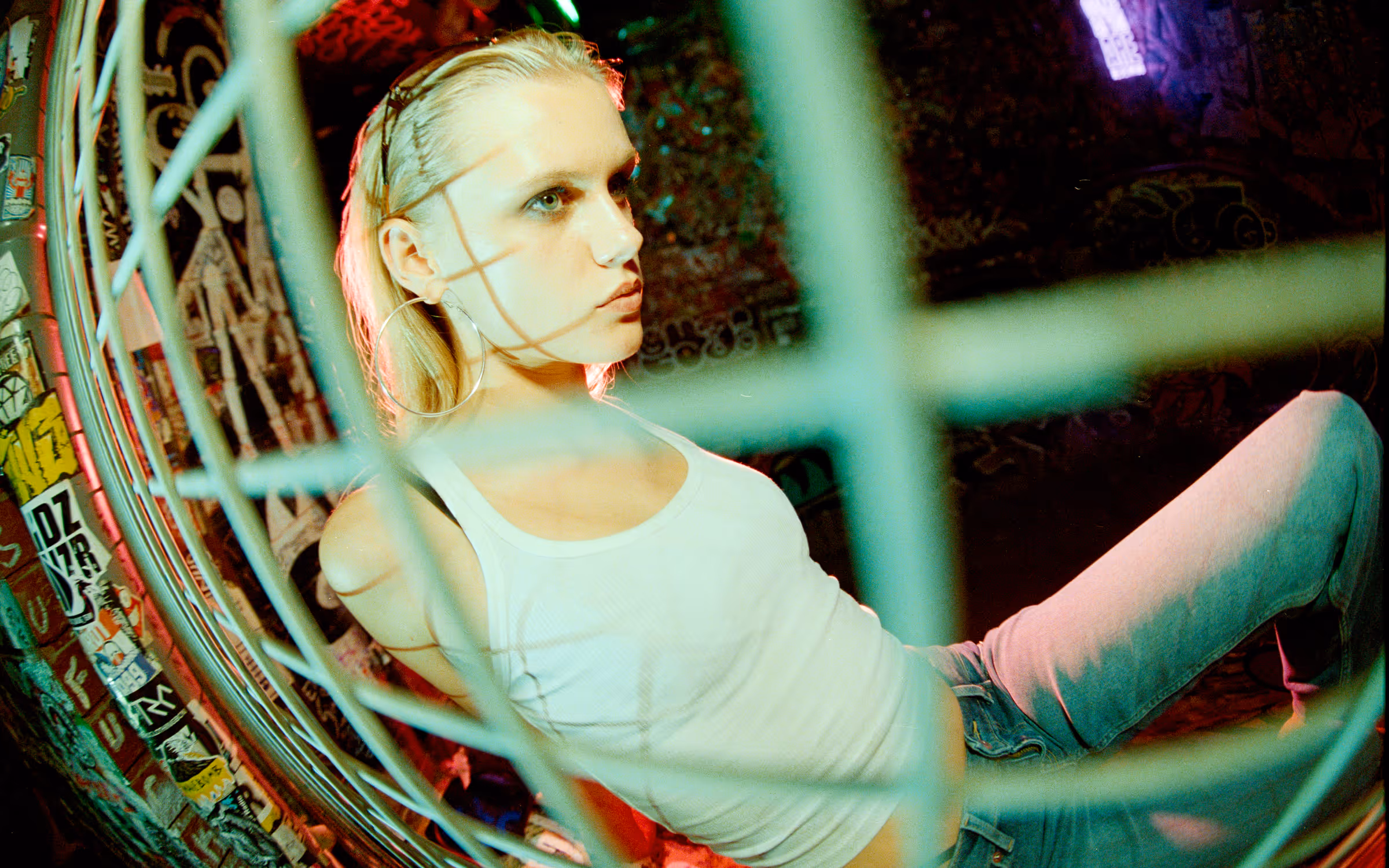 Young person with blonde hair, wearing a white tank top and light blue jeans, sits leaning back inside a metal structure with graffiti-covered walls in the background.