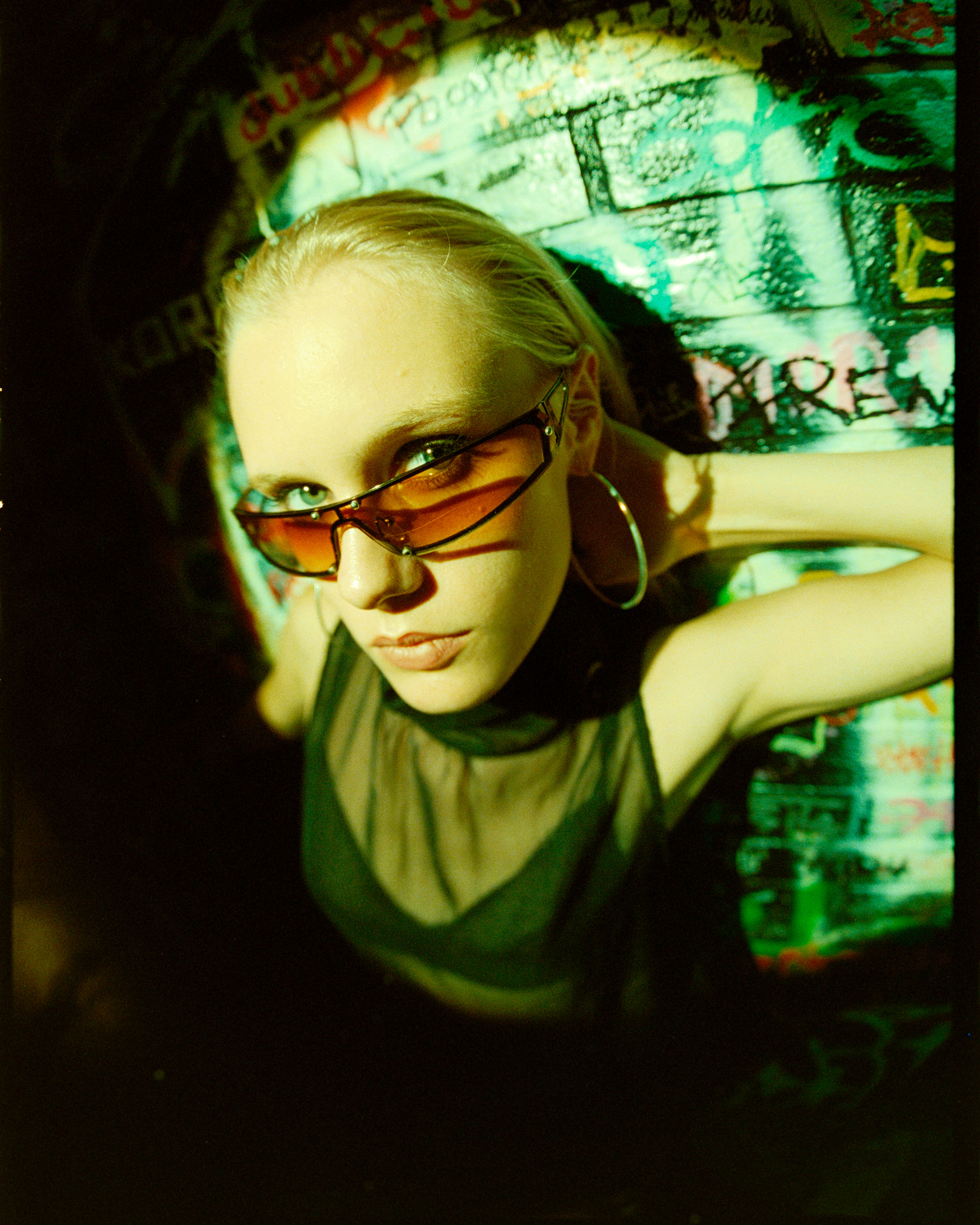 Young woman with slicked-back blonde hair wearing orange-tinted sunglasses and a sheer black top against a colorful graffiti wall.